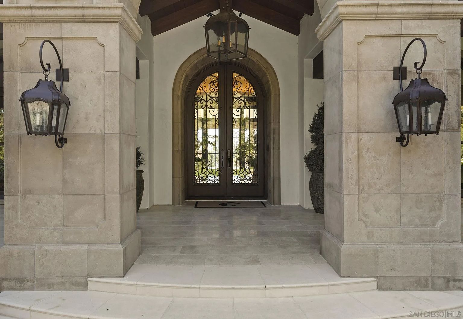 This is a grand entryway featuring a set of arched double doors with intricate ironwork. The entrance is flanked by substantial stone pillars with sconce lighting, leading up to a tiled landing. The overall impression is one of luxury and architectural detail, creating a welcoming and impressive entrance to the property.