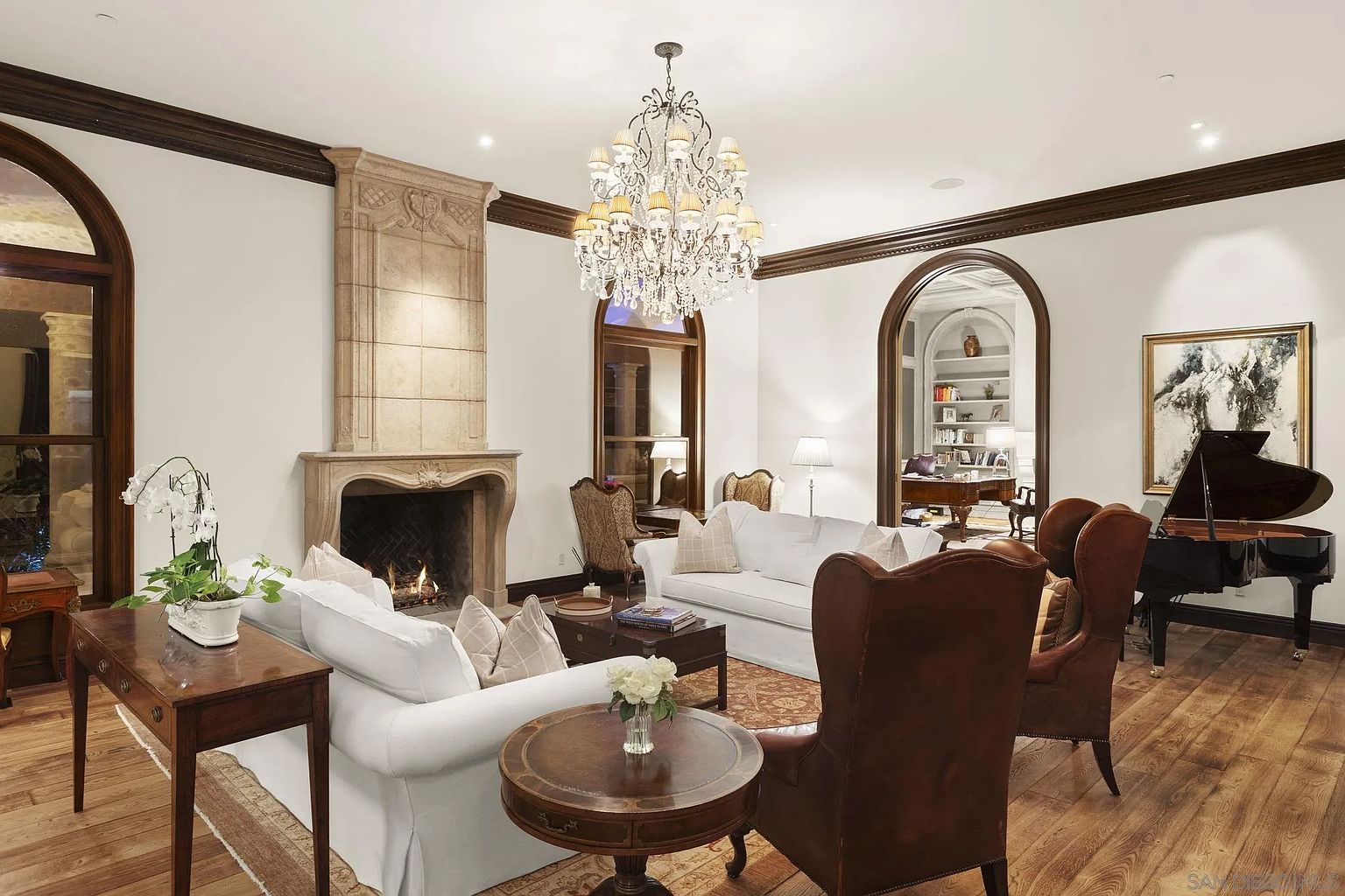 This is an elegant living room featuring a stone fireplace, two white sofas, and two brown leather armchairs. A grand piano sits in the corner, and a large chandelier hangs from the ceiling. The room has hardwood floors and dark wood trim, creating a luxurious and inviting atmosphere.
