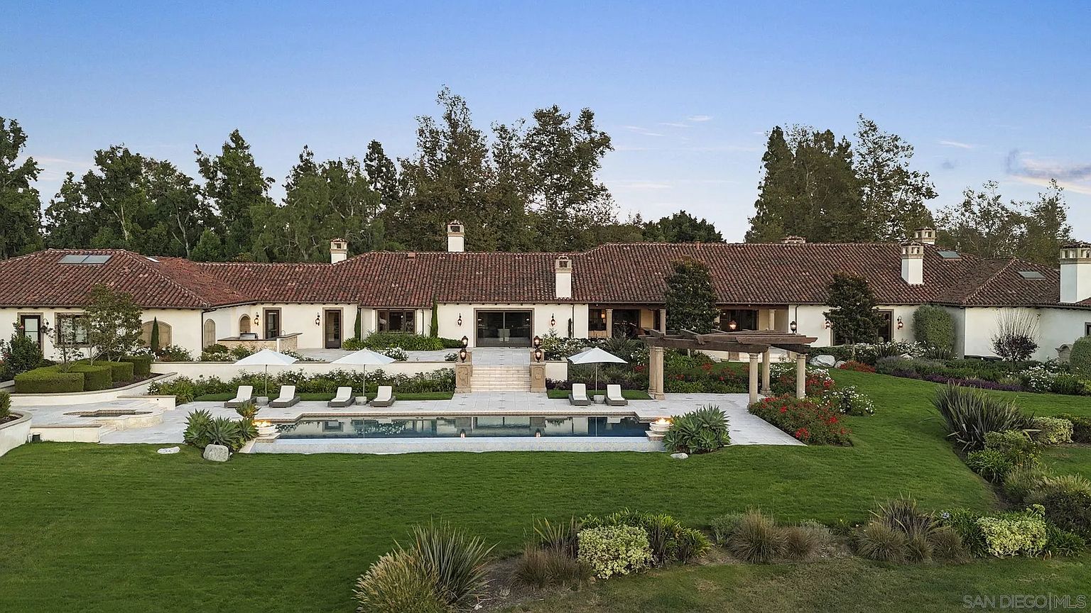 This is a rear view of a luxurious single-story home featuring a red tile roof, a sprawling lawn, and a pristine pool area. The property includes multiple lounge chairs, umbrellas, and a pergola, creating an inviting outdoor living space. The architecture is elegant and timeless, complemented by lush landscaping and mature trees.