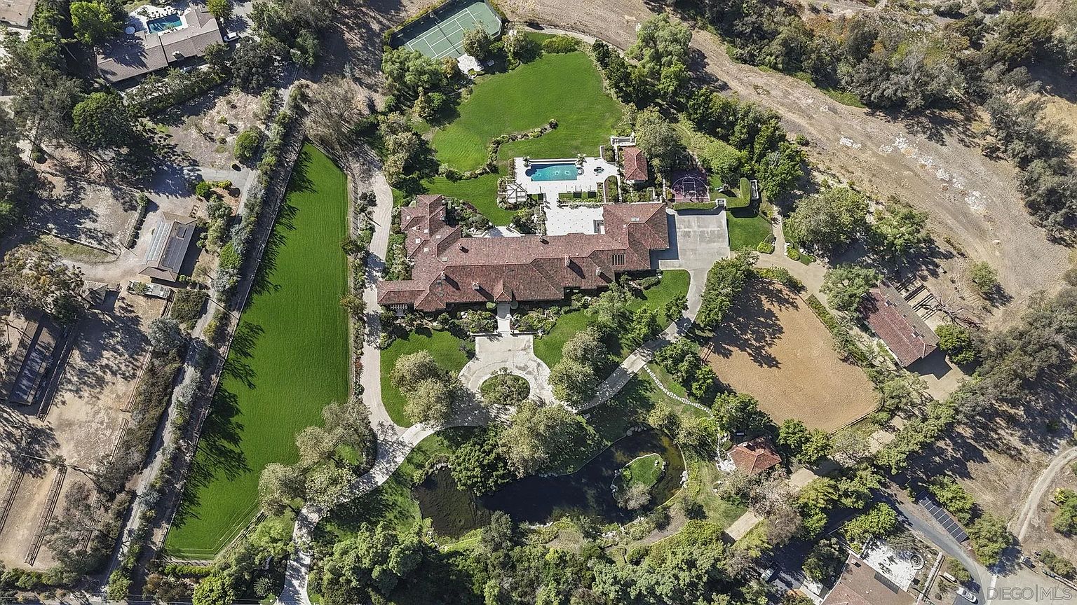 This aerial view showcases an expansive estate featuring a large, luxurious home with a red tile roof, surrounded by lush green lawns, mature trees, and a serene pond. Amenities include a swimming pool, tennis court, and horse riding area, highlighting the property's exceptional outdoor living spaces and privacy. The overall impression is one of grandeur and tranquility.