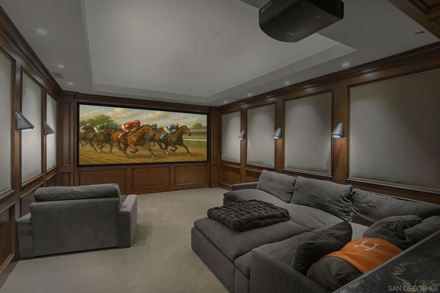 This is an interior shot of a home theater room. The room features wood paneling, sconce lighting, and a large screen displaying a horse racing scene. A large sectional sofa and armchair provide ample seating for viewing, creating a comfortable and luxurious entertainment space.