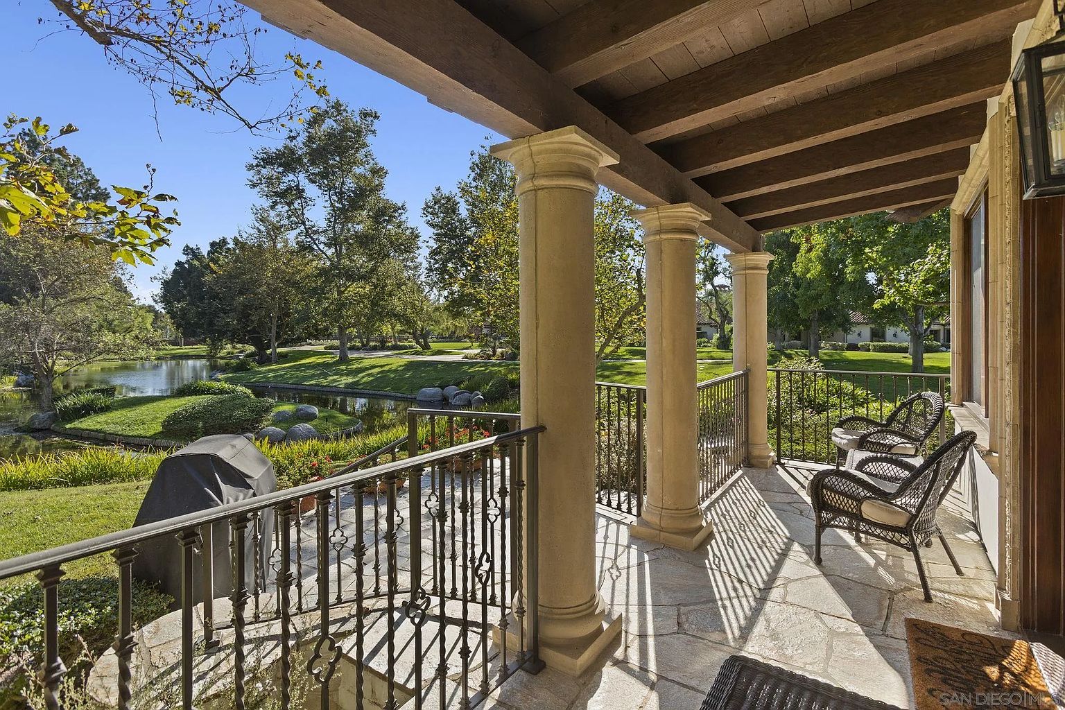 This image showcases a beautiful outdoor patio or balcony area with stone flooring and elegant columns supporting a wooden roof structure. Wrought iron railings provide safety and style, while comfortable wicker chairs invite relaxation. The view overlooks a serene pond and lush greenery, creating a tranquil and inviting atmosphere.