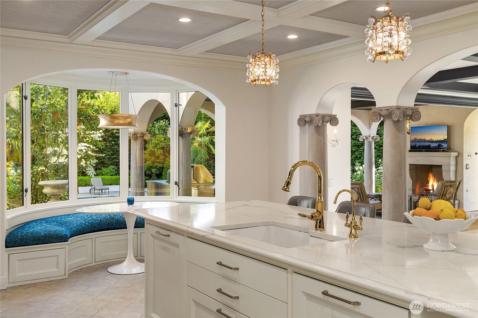 This elegant kitchen features a large central island with a stone countertop and a farmhouse-style sink, complemented by gold-toned fixtures. To the left, a curved breakfast nook with a built-in bench seat sits beneath a series of large windows that look out onto a lush garden. The space is defined by arched architectural openings, decorative columns, and intricate coffered ceilings, creating a sophisticated and airy atmosphere that flows into an adjacent living area with a fireplace.