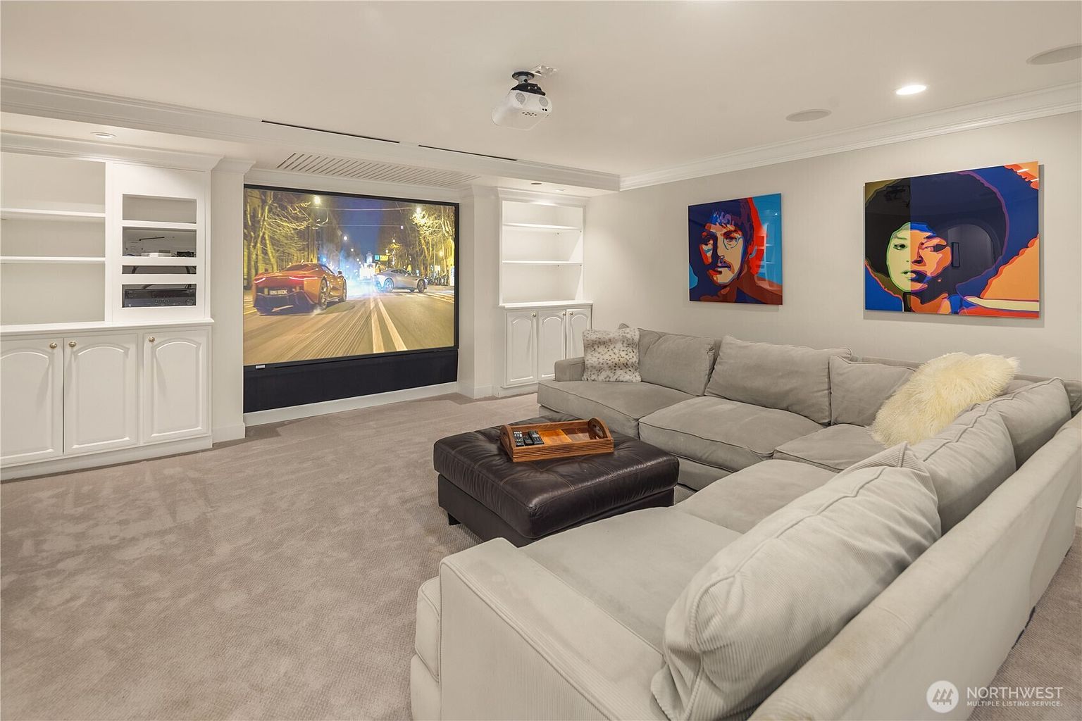 This finished basement serves as a dedicated home theater, featuring a large projection screen flanked by custom white built-in shelving and cabinetry. A spacious, light-gray sectional sofa with a leather ottoman provides comfortable seating, while vibrant pop-art portraits add a modern aesthetic to the neutral walls. The room is illuminated by recessed lighting and includes a ceiling-mounted projector, creating an immersive entertainment environment.