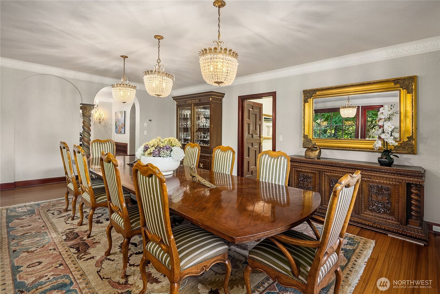 This formal dining room features a large, polished wood table surrounded by chairs with striped upholstery, set atop a traditional patterned area rug. The space is illuminated by three ornate crystal chandeliers and includes a dark wood china cabinet and a matching sideboard topped with a large, gold-framed mirror. The room is characterized by its classic, elegant aesthetic, highlighted by arched architectural details and warm, neutral wall tones.