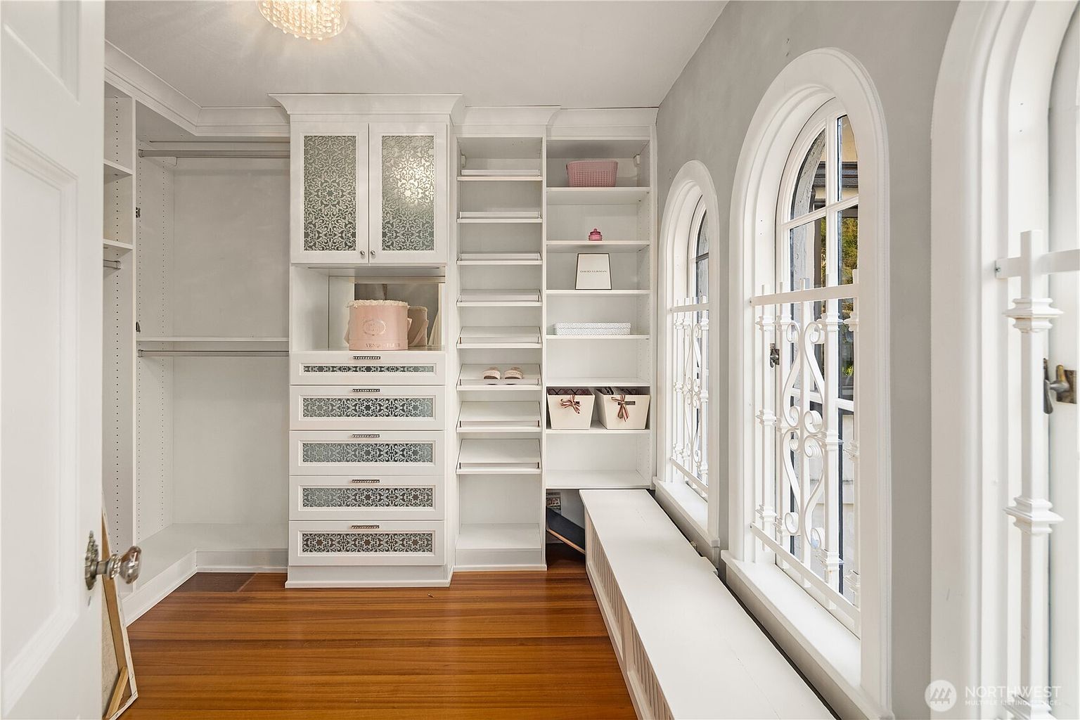 This elegant walk-in closet features custom white cabinetry with decorative patterned glass inserts and integrated shelving. A long, built-in bench runs beneath a series of tall, arched windows that allow ample natural light to fill the space. The room is finished with warm-toned wood flooring and a classic chandelier, creating a sophisticated and organized dressing area.