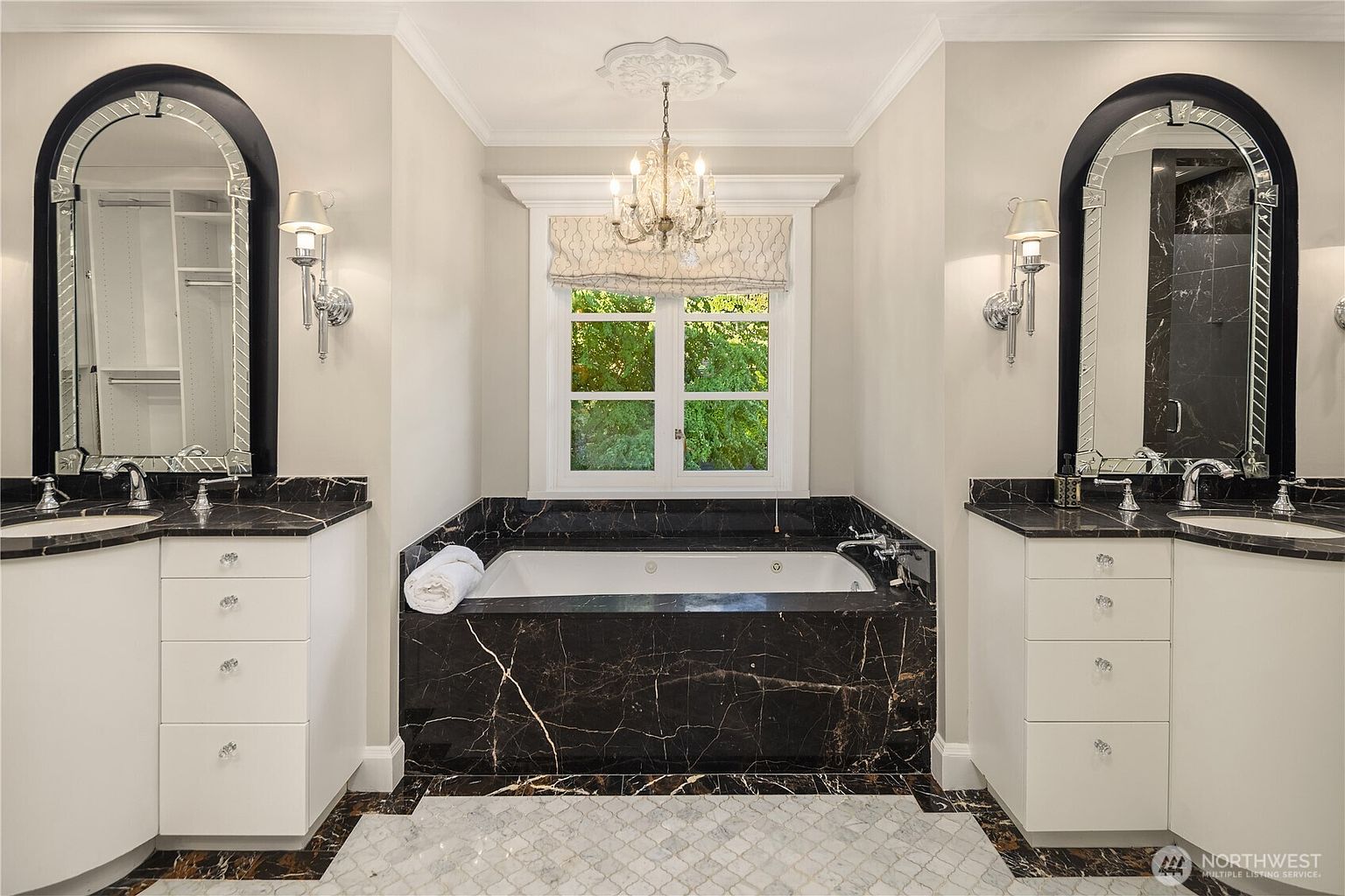 This luxurious primary bathroom features a symmetrical layout with dual vanities flanking a central soaking tub set against a window. The space is defined by dark, veined stone surfaces on the tub surround and vanity countertops, contrasted by crisp white cabinetry and light-toned walls. A crystal chandelier hangs from a decorative ceiling medallion, adding an elegant touch to the sophisticated, high-contrast design.