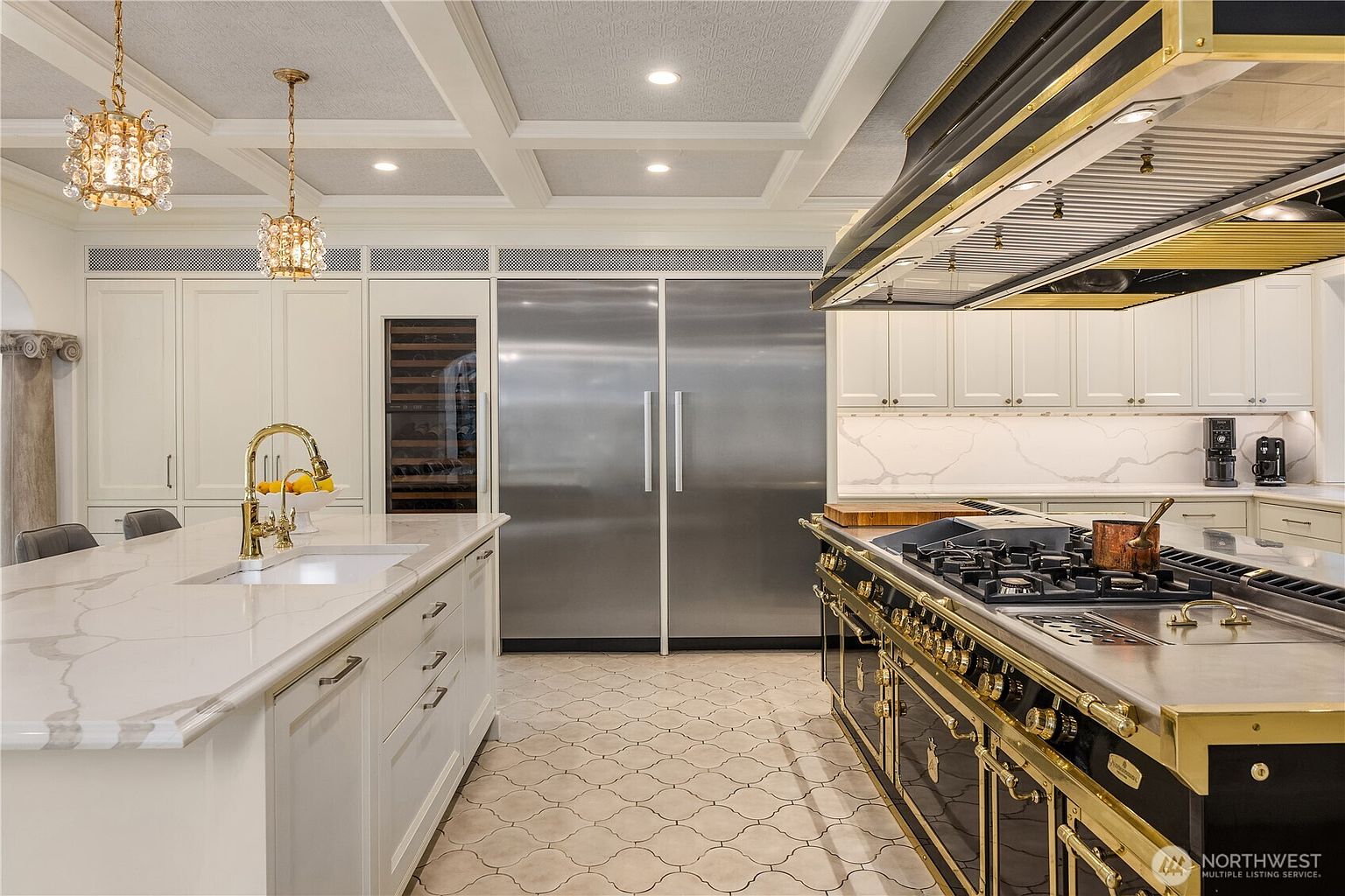 This luxurious kitchen features a large central island with a stone countertop and a gold-toned faucet, complemented by a professional-grade black and gold range with a matching oversized hood. The space is anchored by a massive stainless steel refrigerator and a built-in wine cooler, all set against crisp white cabinetry and a patterned tile floor. The coffered ceiling with recessed lighting and elegant crystal pendant lights adds a sophisticated, high-end aesthetic to the room.