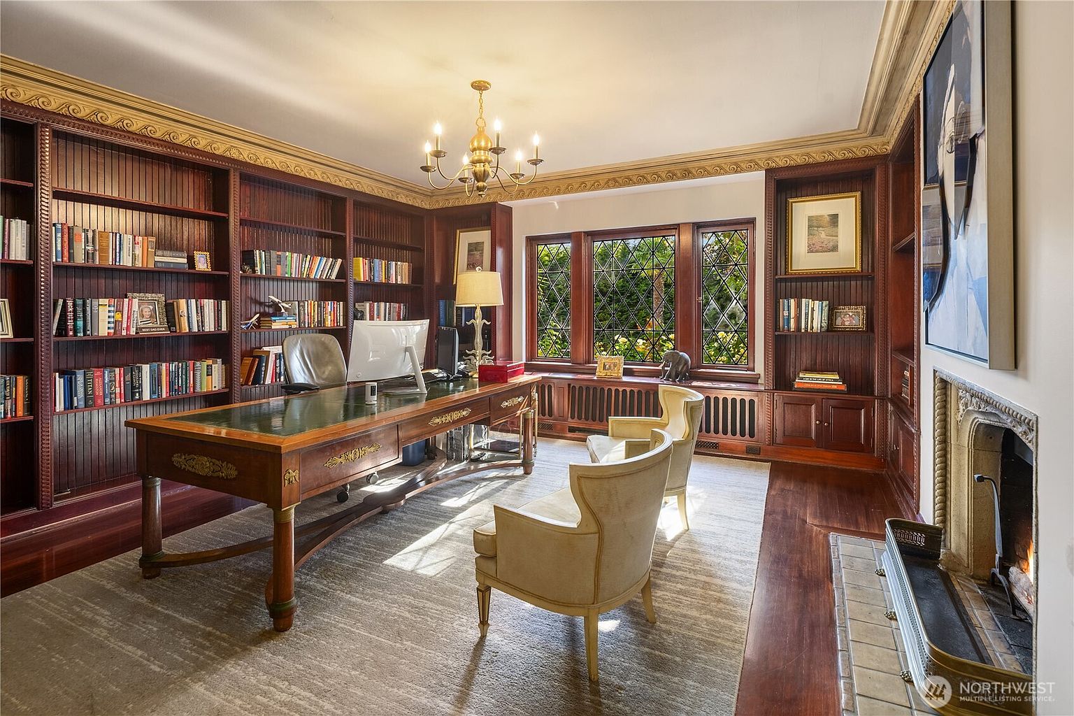 This sophisticated home office features extensive dark wood built-in shelving that spans the walls, creating a classic, scholarly atmosphere. A large, leather-topped executive desk sits centrally on a textured area rug, complemented by two cream-colored armchairs and a fireplace with a stone surround on the right. The room is illuminated by a traditional chandelier and natural light streaming through a series of leaded glass windows.