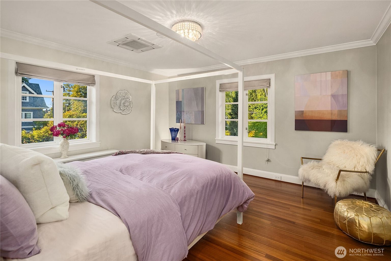 This bright and airy primary bedroom features a white canopy bed frame as the central focal point, dressed in soft lavender bedding. The room is accented by light gray walls, warm wood flooring, and two large windows that provide ample natural light and views of the surrounding greenery. A plush, furry accent chair and a gold-toned ottoman add a touch of texture and luxury to the space, while abstract wall art completes the sophisticated aesthetic.