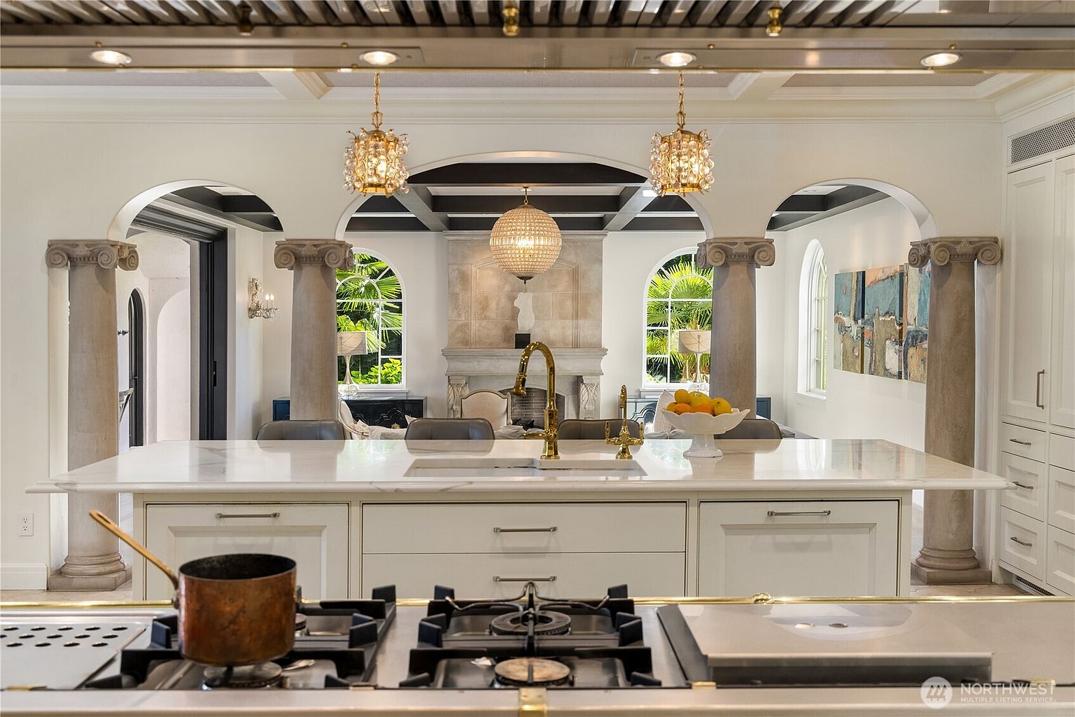 This elegant kitchen features a large central island with a stone countertop, gold-toned fixtures, and a prominent gas cooktop in the foreground. The space is defined by classical architectural elements, including decorative columns that frame an open view into a living area with a stone-clad chimney breast and arched windows. The design aesthetic is sophisticated and grand, utilizing a neutral color palette and ornate lighting fixtures to create a luxurious atmosphere.