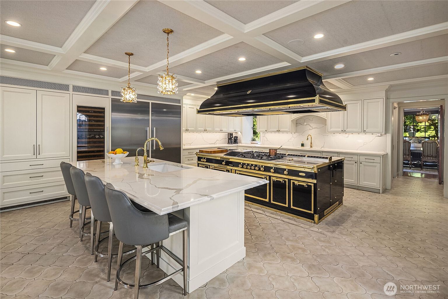 This luxurious, high-end kitchen features a large central island with a stone countertop and seating for three, complemented by a professional-grade black and gold range with a matching oversized hood. The space is defined by its coffered ceiling, light-colored cabinetry, and intricate patterned tile flooring, creating an elegant and sophisticated atmosphere. A view into an adjacent dining area is visible through the doorway, adding depth to the perspective of this spacious culinary environment.