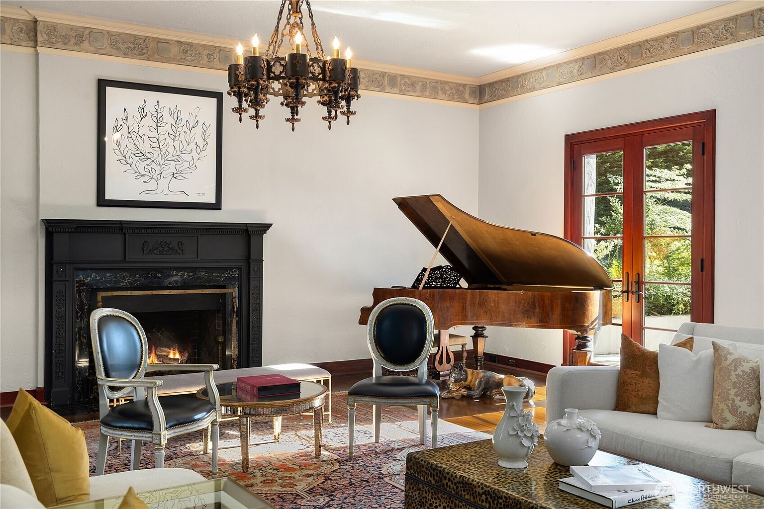 This elegant living room features a grand piano as a focal point, complemented by a sophisticated black fireplace surround and a decorative crown molding border. The space is furnished with classic chairs, a plush sofa, and a patterned area rug, all illuminated by a traditional chandelier. Large French doors provide a view of the outdoors, creating a refined and inviting atmosphere.