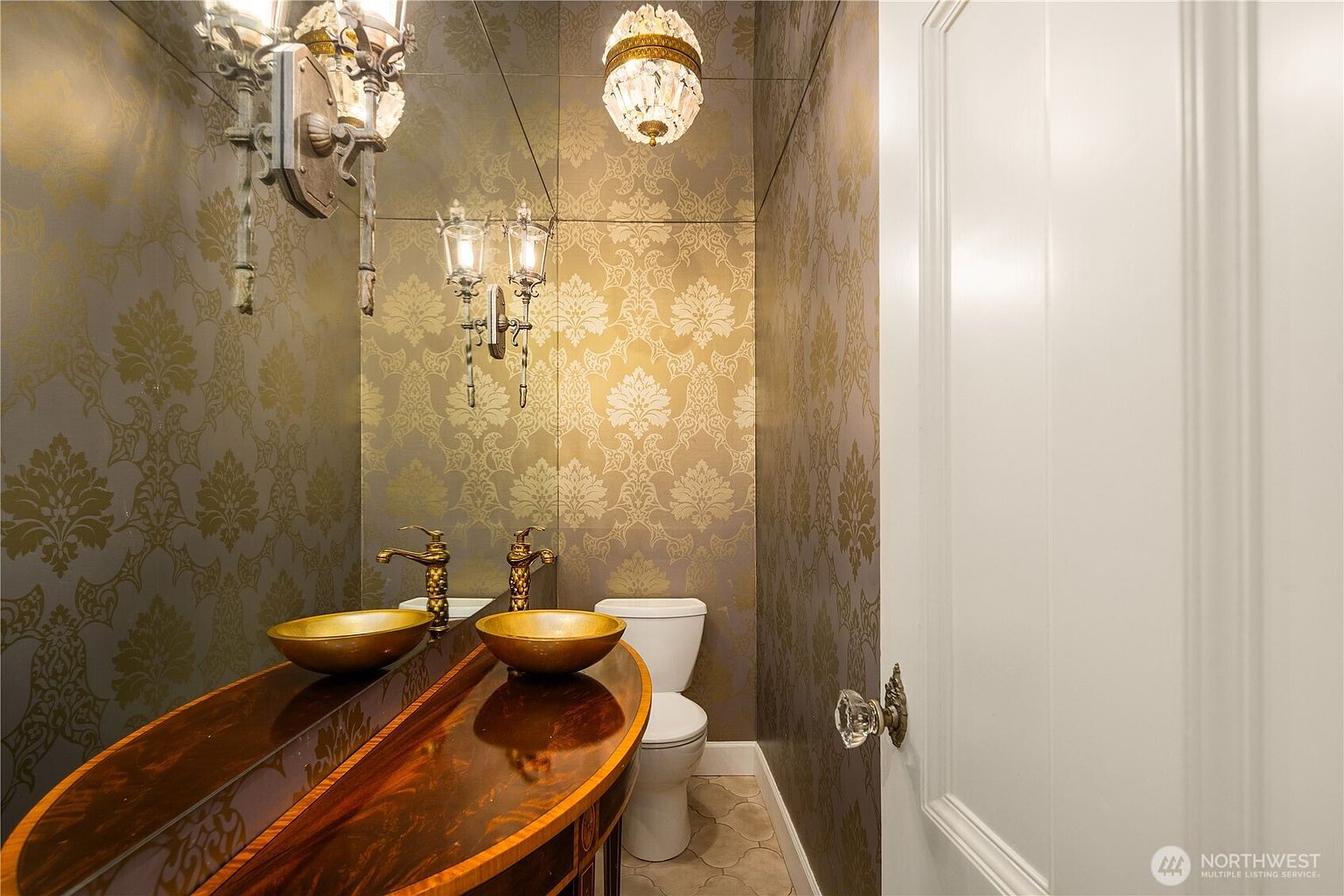 This elegant powder room features ornate damask-patterned wallpaper and a mirrored wall that enhances the sense of space. A dark wood vanity with a gold-toned vessel sink and matching faucet adds a touch of luxury, complemented by decorative wall sconces and a crystal-style ceiling fixture. The room is finished with a white toilet and neutral-toned floor tiles, creating a sophisticated and intimate atmosphere.