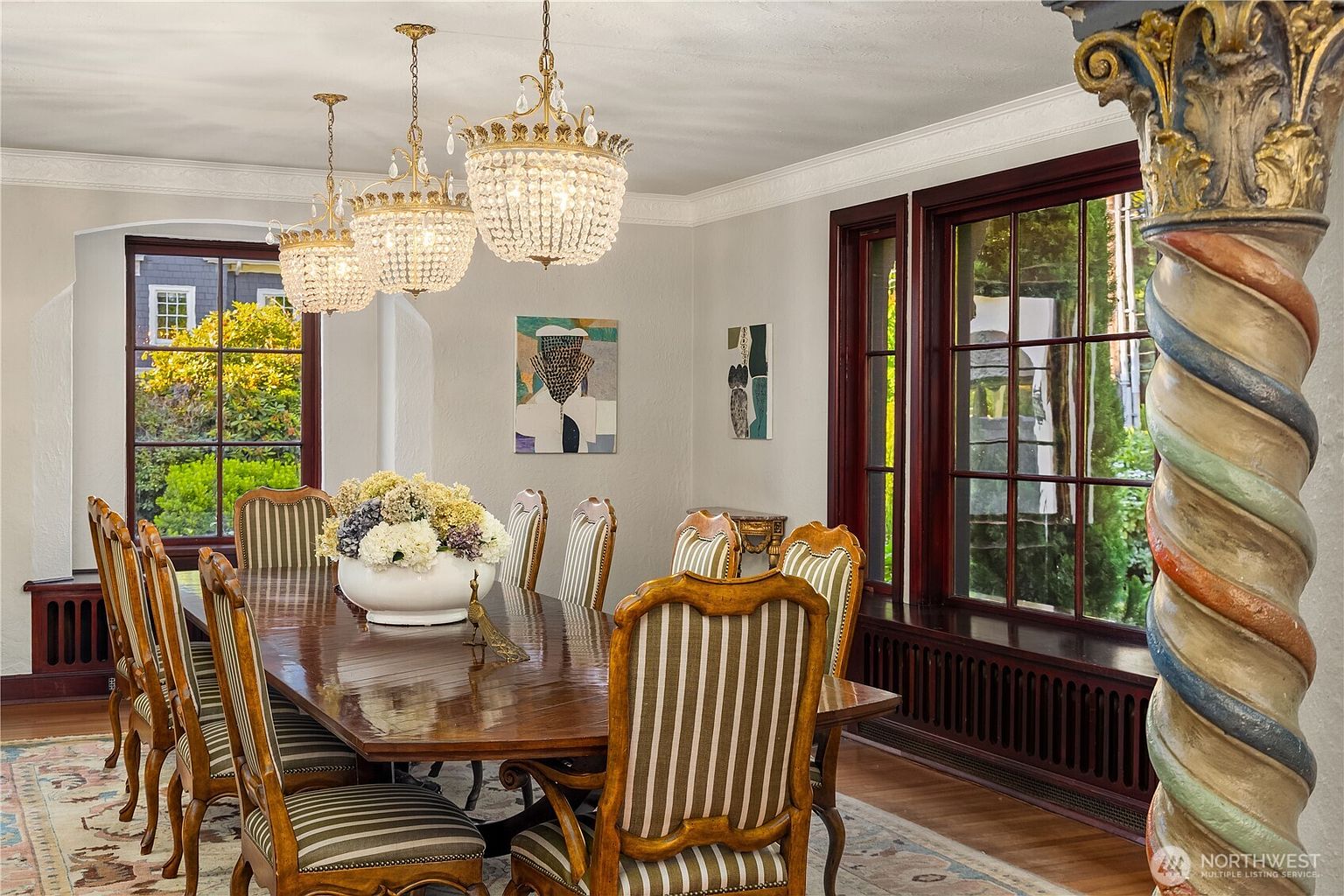 This elegant dining room features a long, polished wood table surrounded by upholstered chairs with striped fabric. Three ornate crystal chandeliers hang above the table, casting a warm glow over the space, while large windows with dark wood frames provide ample natural light. A decorative, spiraled column stands in the foreground, adding a unique architectural detail to the sophisticated, traditional interior.