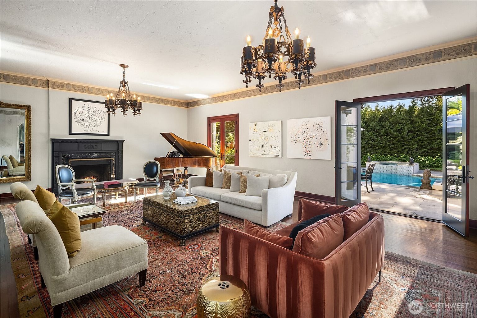 This elegant living room features a grand piano, a dark fireplace surround, and two ornate chandeliers that anchor the space. The room is furnished with a mix of plush seating, including a velvet sofa and a patterned ottoman, all arranged on a large, intricate area rug. Large glass doors open directly to a patio and pool area, creating a seamless transition between the sophisticated interior and the outdoor living space.
