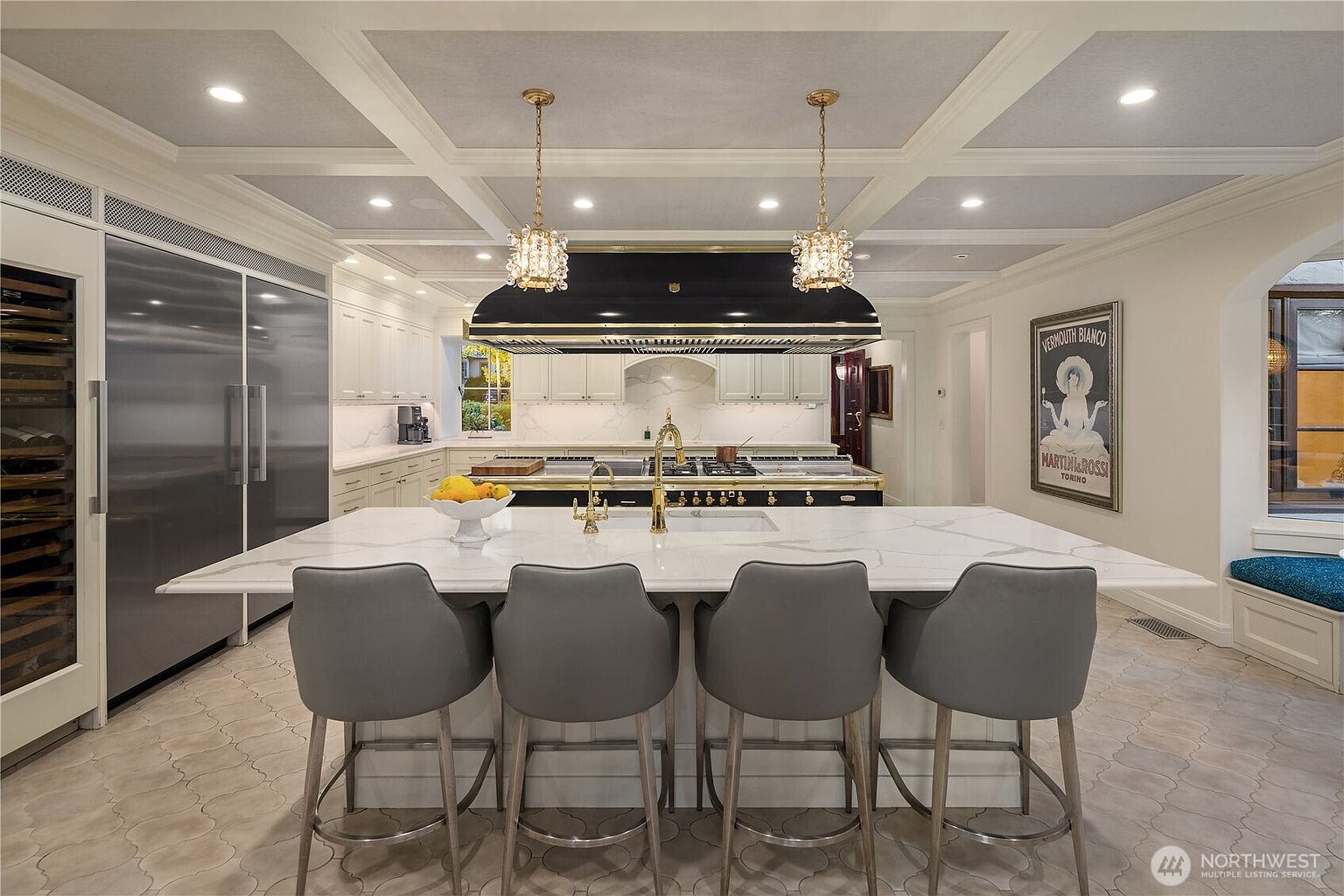 This sophisticated kitchen features a large central island with a white stone countertop and four grey upholstered bar stools. A prominent black and gold range hood hangs above a professional-grade stove, complemented by white cabinetry and a patterned tile floor. The space is illuminated by elegant crystal pendant lights and recessed ceiling lighting, creating a luxurious and well-appointed culinary environment.