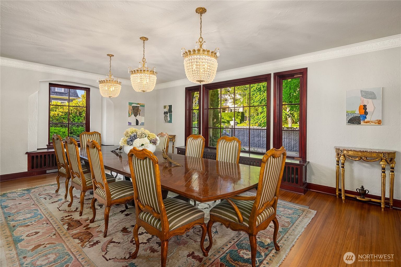 This formal dining room features a large, dark wood table surrounded by eight striped upholstered chairs, set atop a traditional patterned area rug. Three ornate crystal chandeliers hang above the table, providing elegant lighting, while large windows with dark wood frames offer views of the lush greenery outside. A decorative console table with a stone top sits against the wall, complemented by framed artwork, creating a sophisticated and classic atmosphere.