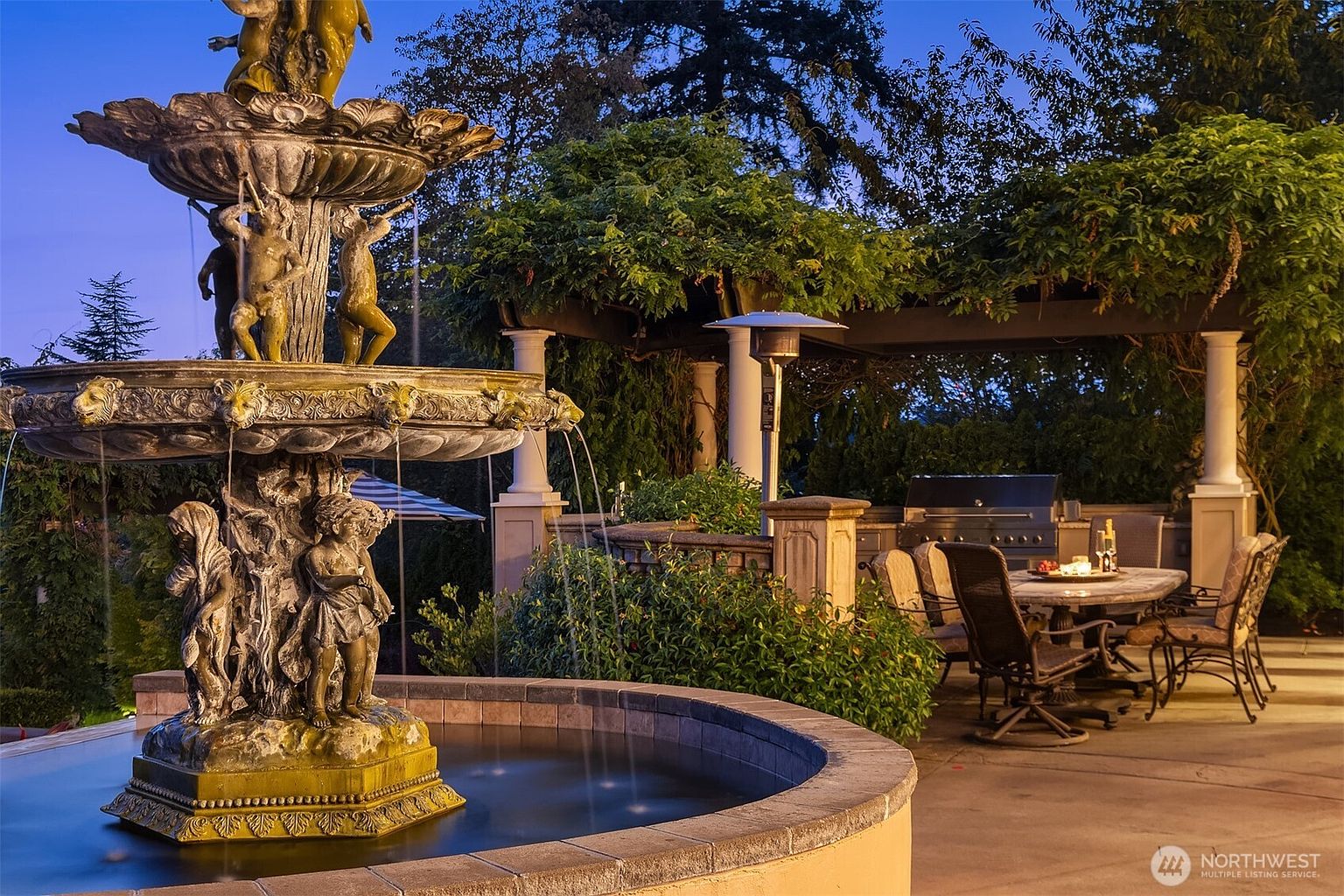This elegant outdoor living space features a grand, multi-tiered stone fountain as the centerpiece, set against a backdrop of lush greenery and a pergola-covered dining area. The patio is furnished with a dining set and a built-in grill, creating a sophisticated atmosphere perfect for evening entertaining. The warm lighting and tranquil water feature provide a luxurious and inviting ambiance for outdoor relaxation.