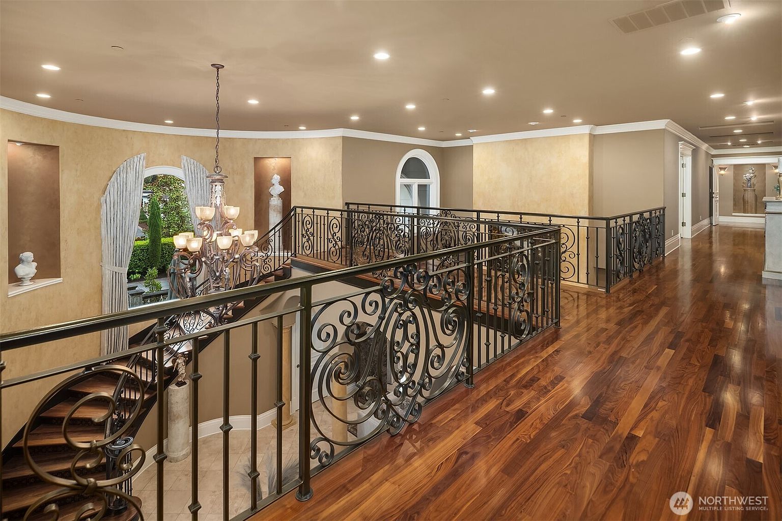 This elegant upper-level landing features rich, polished hardwood flooring and an ornate wrought-iron railing that overlooks the grand staircase below. The space is illuminated by recessed lighting and a sophisticated chandelier, while decorative wall niches house classical busts, enhancing the home's luxurious and traditional aesthetic. The perspective captures the expansive flow of the hallway, emphasizing the high-end craftsmanship and open architectural design.