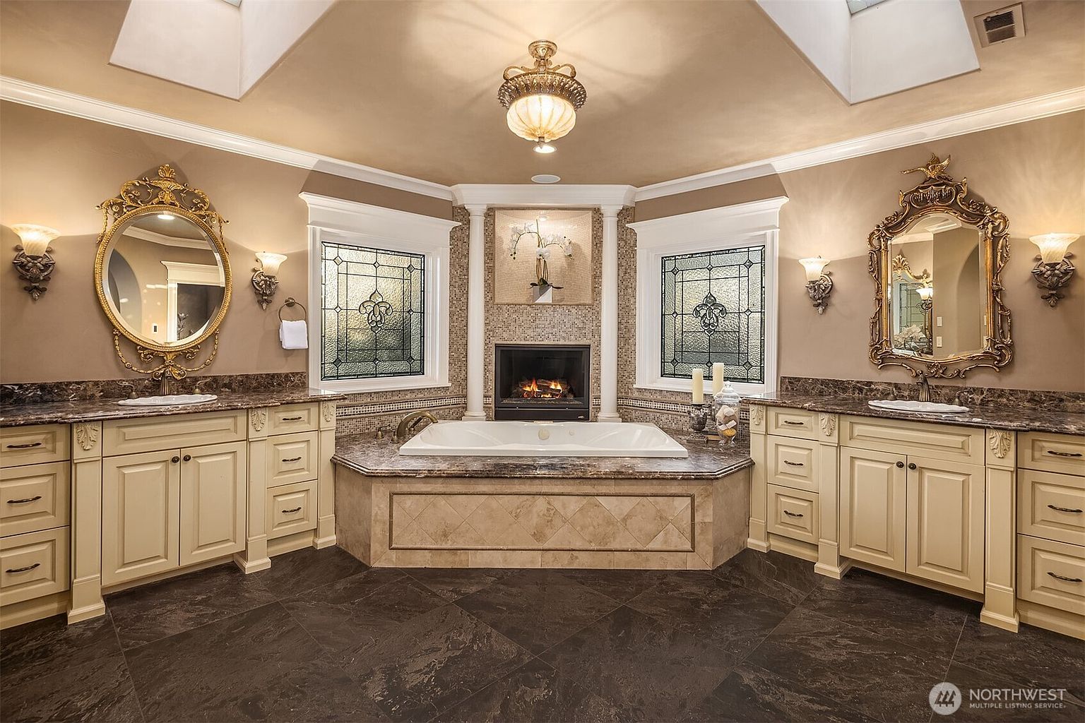 This luxurious primary bathroom features a symmetrical layout with dual vanities flanking a central jetted soaking tub and fireplace. The space is adorned with ornate gold-framed mirrors, stained glass windows, and elegant tile work, creating a sophisticated and opulent atmosphere. The perspective is a wide, centered shot that emphasizes the grand, spa-like design of the room.