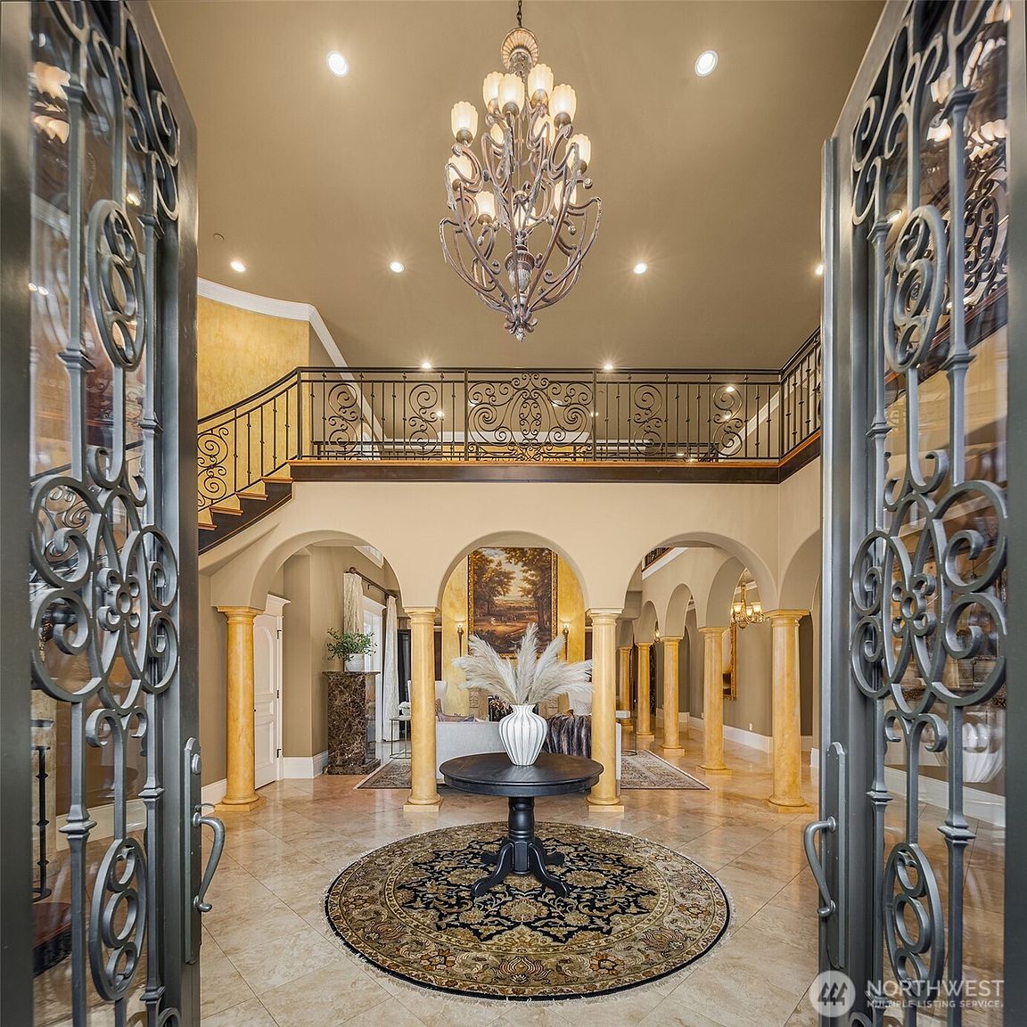 This grand foyer features an impressive two-story entrance with a sweeping staircase and an ornate wrought-iron railing. The space is defined by elegant arched columns, polished stone flooring, and a dramatic chandelier that serves as the centerpiece. The perspective is from the doorway looking inward, showcasing a luxurious, Mediterranean-inspired architectural style.