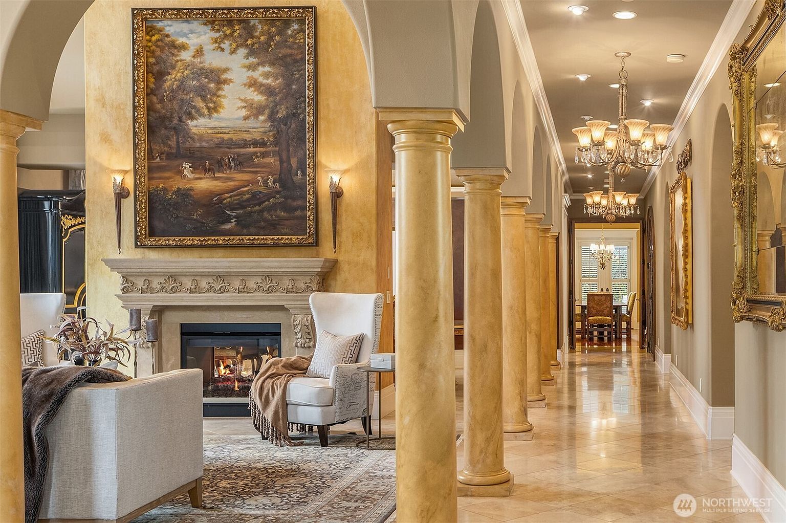 This elegant living room features a grand stone fireplace adorned with a large landscape painting and flanked by classic wall sconces. A series of stately, faux-finished columns creates a dramatic transition into a long, polished-tile hallway that leads toward a formal dining area. The space exudes a sophisticated, Mediterranean-inspired luxury with warm, golden tones and high-end architectural detailing.