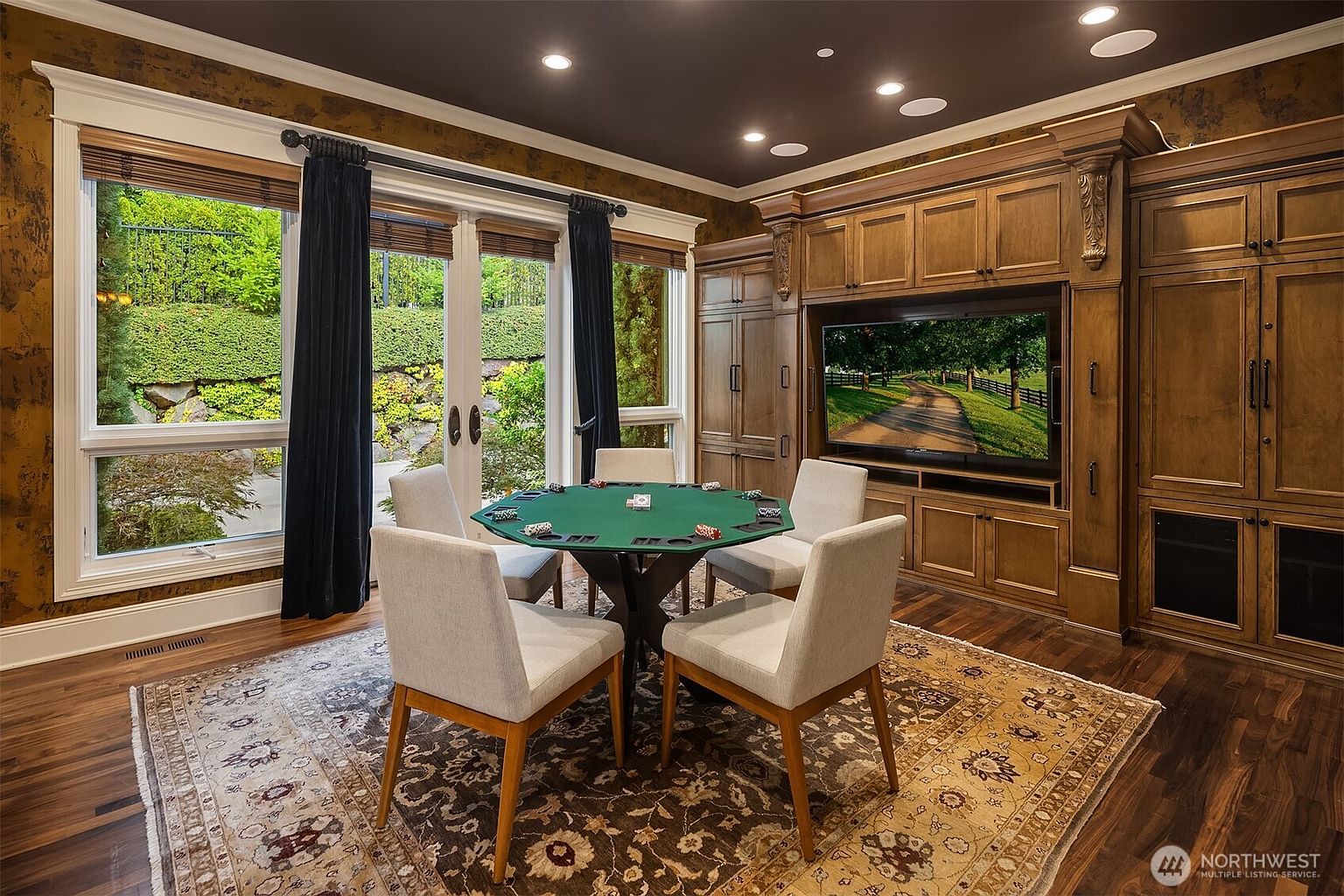 This sophisticated game room or study features a central poker table surrounded by four upholstered chairs, set atop an ornate, traditional-style area rug. The room is anchored by a large, custom-built wooden entertainment center with integrated cabinetry and a television, while floor-to-ceiling windows with dark curtains provide a view of the lush outdoor landscaping. The dark ceiling with recessed lighting and warm-toned walls create an intimate, high-end atmosphere perfect for leisure or focused work.