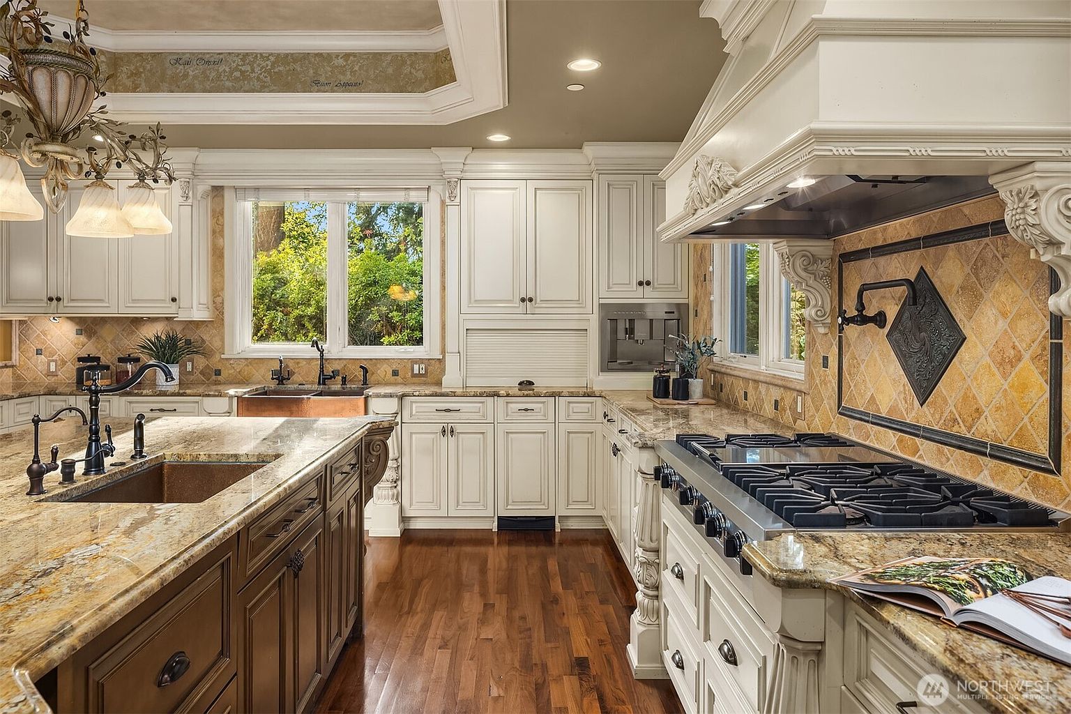 This elegant, traditional-style kitchen features custom off-white cabinetry, rich granite countertops, and a large central island with a dark wood finish. The space is equipped with high-end stainless steel appliances, including a professional-grade gas range with a decorative hood and a built-in coffee system. A warm, inviting atmosphere is created by the combination of hardwood flooring, intricate crown molding, and natural light streaming through the windows.