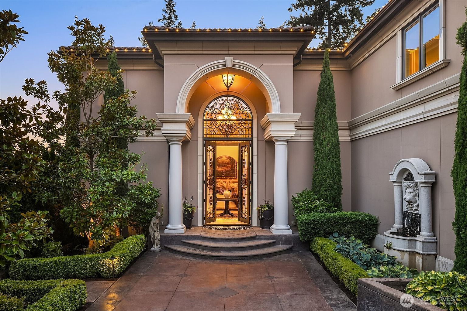 This elegant front entryway features a grand arched doorway framed by classic white columns and intricate wrought-iron detailing. The scene is enhanced by manicured hedges, tall cypress trees, and a stone wall fountain, creating a sophisticated and welcoming atmosphere. The warm lighting from within and the soft evening glow highlight the architectural craftsmanship of this luxury home.