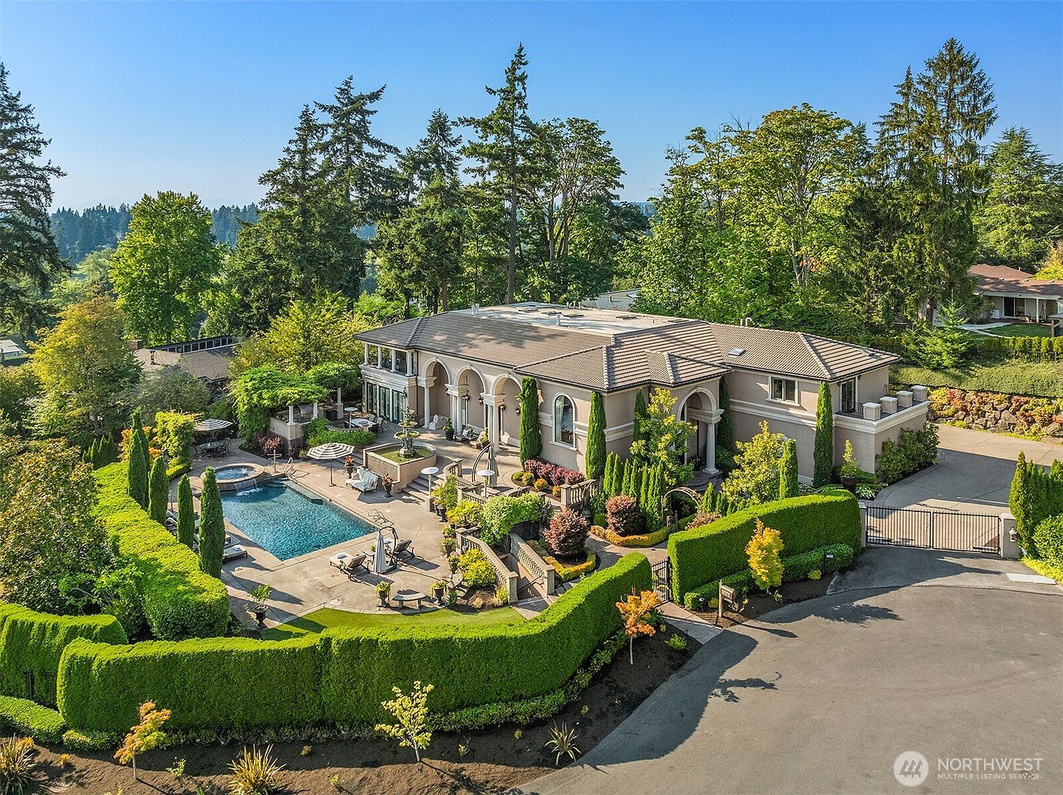 This high-angle aerial view captures a luxurious Mediterranean-style estate nestled within a lush, tree-lined landscape. The property features a sprawling residence with arched architectural details, a pristine swimming pool with an integrated spa, and meticulously manicured hedges that define the outdoor living spaces. The scene conveys an atmosphere of exclusivity and refined elegance, highlighting the seamless integration of the home with its private, park-like surroundings.