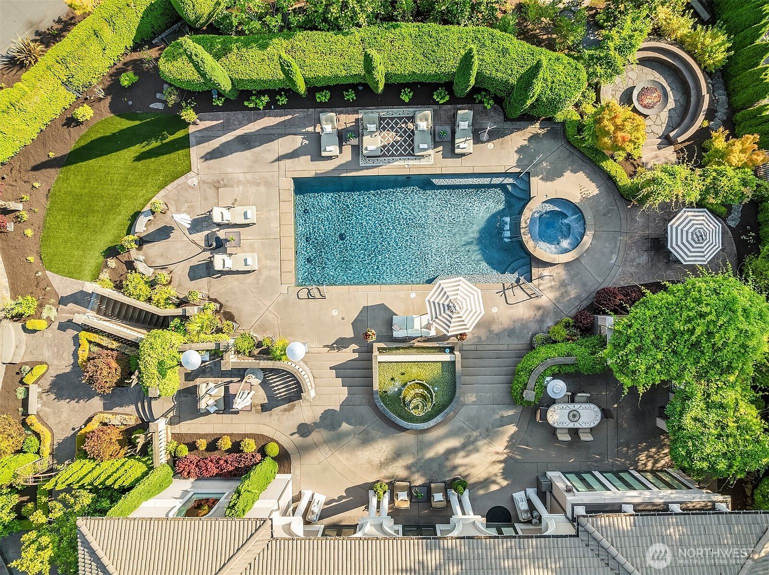This high-angle aerial view captures a luxurious backyard oasis featuring a large rectangular swimming pool with an integrated circular spa. The expansive concrete patio is meticulously landscaped with manicured hedges, a fire pit area, a water feature, and multiple seating arrangements, creating a resort-like atmosphere. The perspective highlights the seamless integration of the home's architecture with its sophisticated outdoor living spaces.