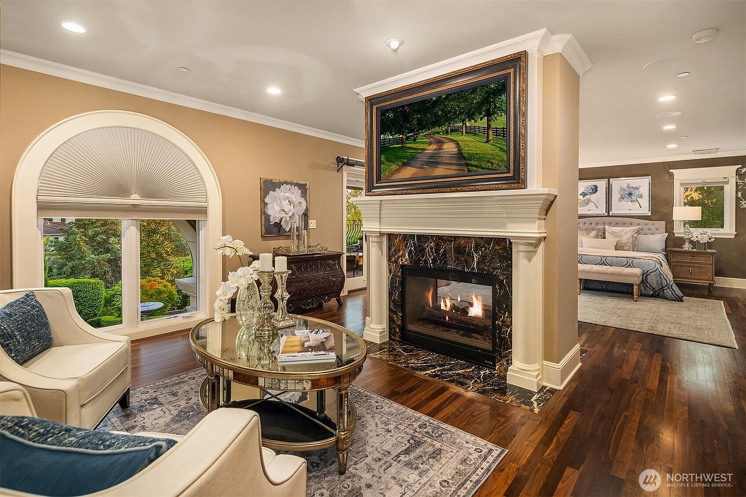 This elegant living room features a sophisticated open-concept design with a central fireplace serving as a focal point, topped by a large framed television. The space is anchored by a plush area rug, a mirrored coffee table, and comfortable cream-colored armchairs, while a seamless transition leads into a cozy bedroom visible in the background. Large arched windows flood the room with natural light, highlighting the rich hardwood floors and refined, traditional decor.