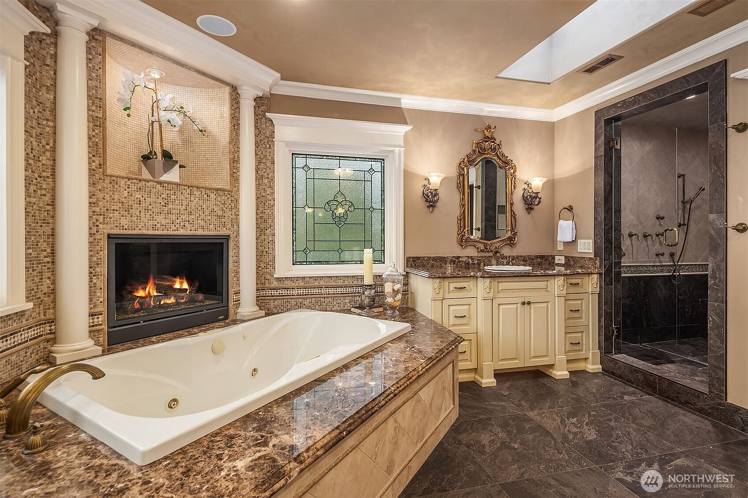 This luxurious primary bathroom features a grand jetted soaking tub set against a fireplace, creating a spa-like atmosphere. The space is adorned with rich marble countertops, ornate gold-framed mirrors, and a stained-glass window, all complemented by a spacious walk-in shower with dark stone tiling. The overall aesthetic is opulent and sophisticated, blending classic architectural details with high-end finishes.