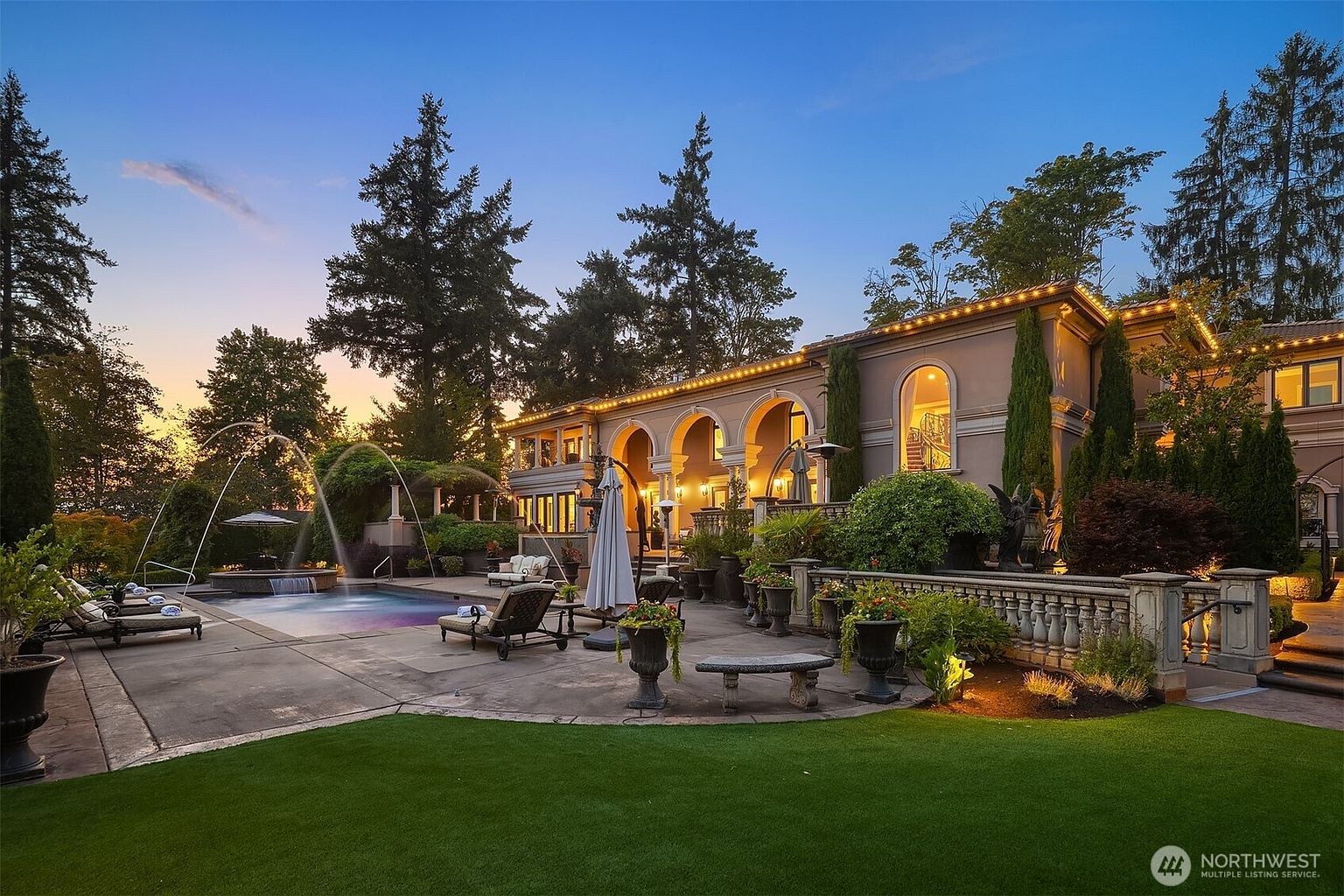 This stunning rear exterior view captures a luxurious Mediterranean-style estate at dusk, featuring a sprawling patio, a custom swimming pool with integrated fountains, and elegant arched architectural details. The meticulously landscaped grounds, illuminated by warm accent lighting, create an inviting and sophisticated atmosphere perfect for outdoor entertaining. The perspective highlights the seamless integration between the grand residence and its resort-like backyard amenities.