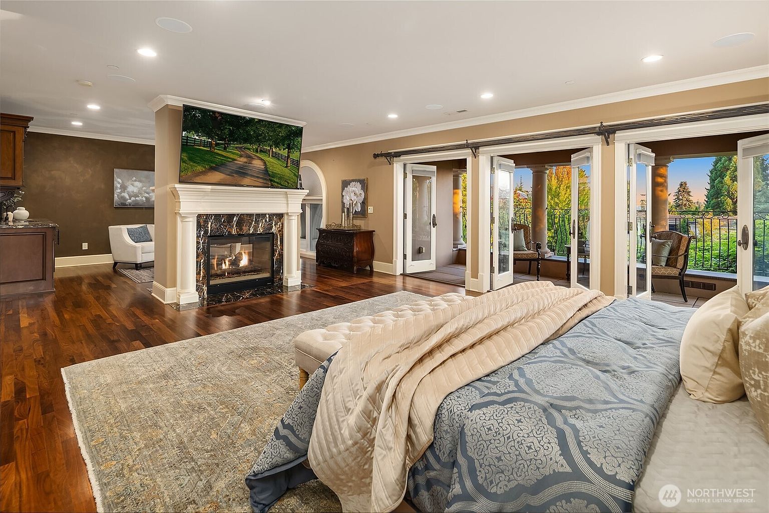 This luxurious primary bedroom features a grand fireplace with a dark marble surround and a mounted television, serving as a focal point in the room. The space is anchored by a large, plush bed with patterned bedding and opens onto a private balcony through multiple sets of French doors, allowing for abundant natural light. The rich hardwood floors and warm, neutral wall tones create an elegant and inviting atmosphere.