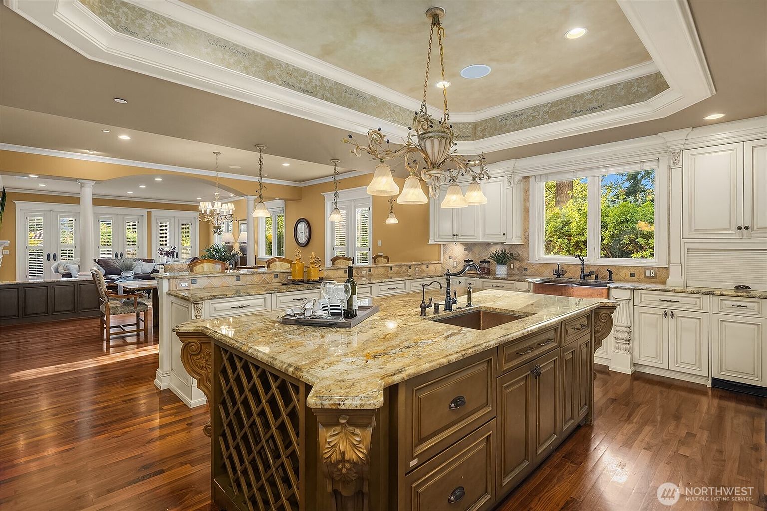 This elegant, traditional-style kitchen features a large, ornate central island with a granite countertop and built-in wine storage, complemented by rich hardwood flooring. The space is highlighted by custom white cabinetry, a decorative tray ceiling with intricate molding, and a grand chandelier that serves as a focal point. The open-concept design flows seamlessly into an adjacent living area, creating a warm and inviting atmosphere perfect for entertaining.