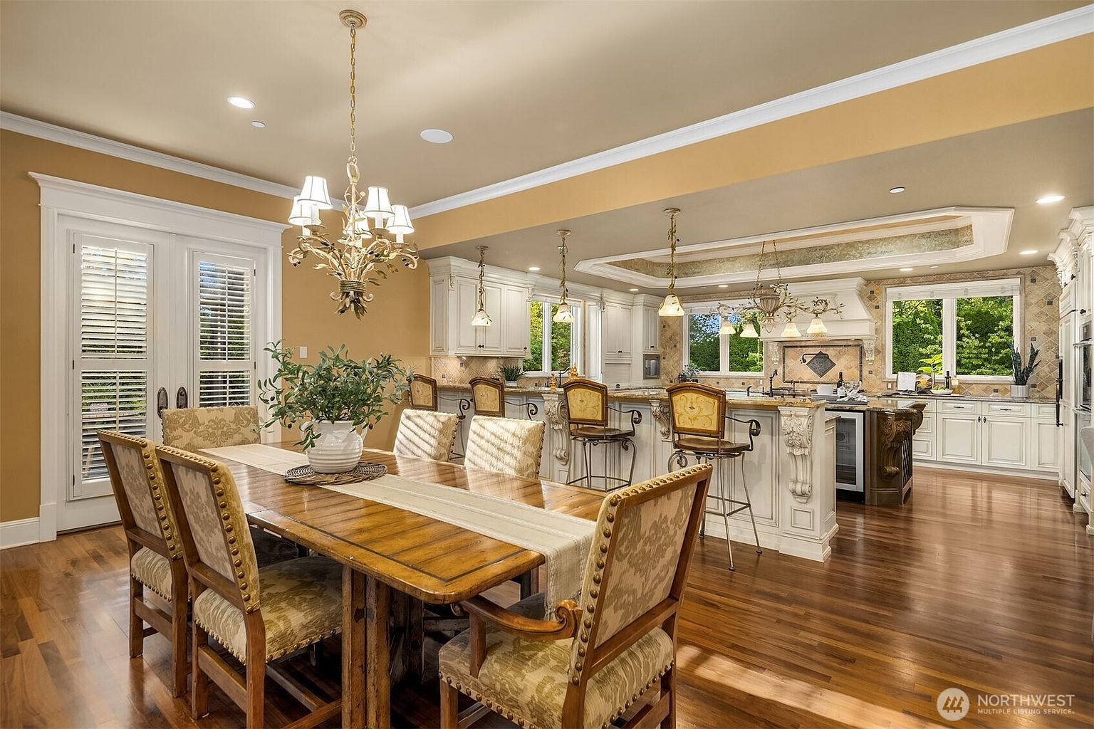 This expansive open-concept kitchen and dining area features a warm, traditional aesthetic with rich hardwood flooring and custom white cabinetry. A large wooden dining table sits in the foreground, while the kitchen island provides ample seating with ornate bar stools and decorative corbels. The space is illuminated by elegant chandeliers and recessed lighting, creating a sophisticated and inviting atmosphere perfect for entertaining.