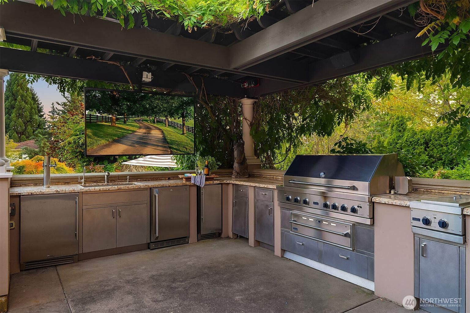 This luxurious outdoor kitchen is nestled under a covered pergola, featuring high-end stainless steel appliances, including a large professional-grade grill and multiple under-counter refrigerators. The space is equipped with a wall-mounted television, making it an ideal setup for outdoor entertainment and dining. The stone countertops and clean, modern cabinetry create a sophisticated atmosphere that seamlessly blends with the lush, green garden backdrop.