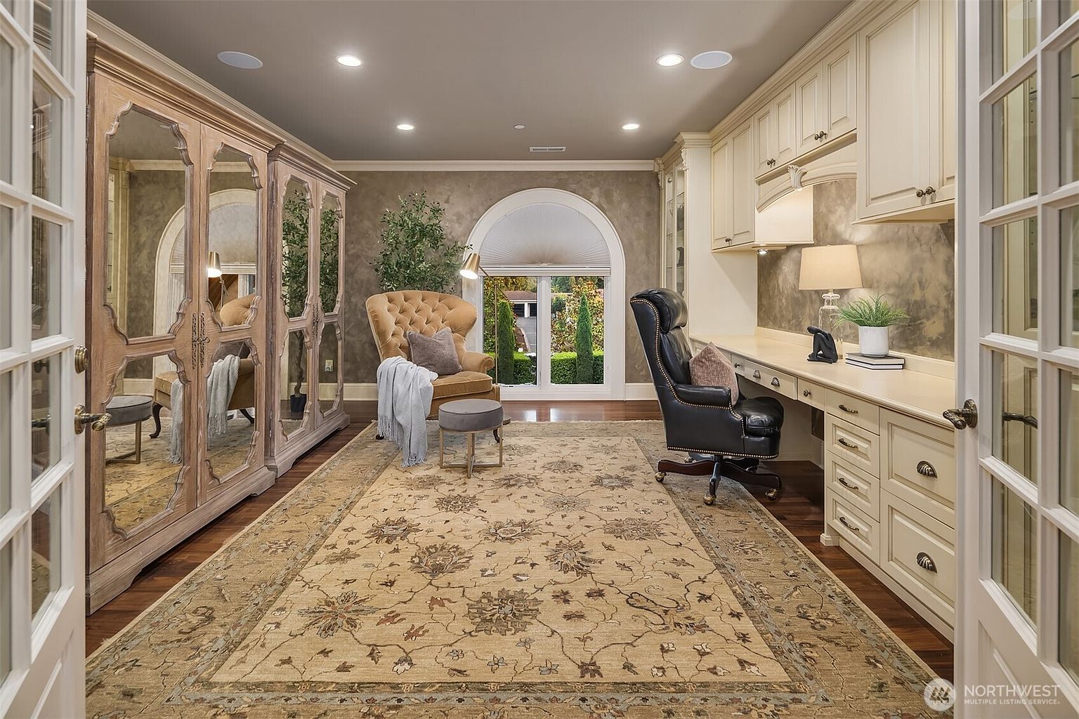 This elegant home office features a sophisticated blend of traditional and classic design elements, including a large, ornate mirrored armoire and built-in cream-colored cabinetry. A plush, tufted wingback chair sits near a large arched window, while a dark leather executive chair is positioned at the expansive desk, all set atop a detailed, traditional-style area rug. The room exudes a sense of refined productivity and luxury, captured from a centered, eye-level perspective that highlights the symmetry and high-end finishes of the space.