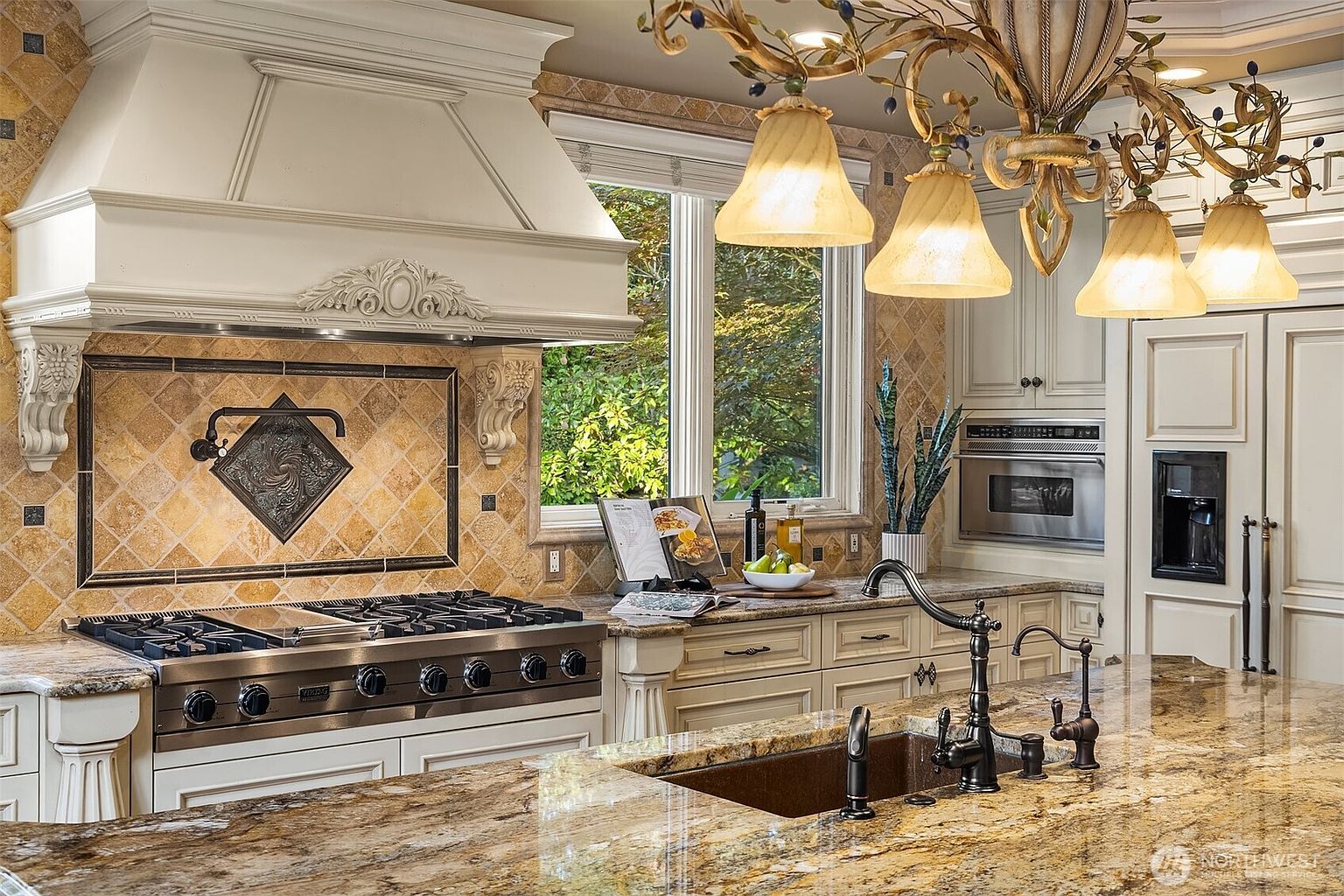 This elegant, high-end kitchen features a large granite-topped island with a dark bronze faucet and a professional-grade Viking range set beneath a custom, ornate white range hood. The backsplash is composed of warm-toned, diamond-patterned stone tiles, complemented by classic cream-colored cabinetry and a decorative wrought-iron chandelier. The perspective is a warm, eye-level shot that highlights the luxurious textures and traditional craftsmanship of the space.