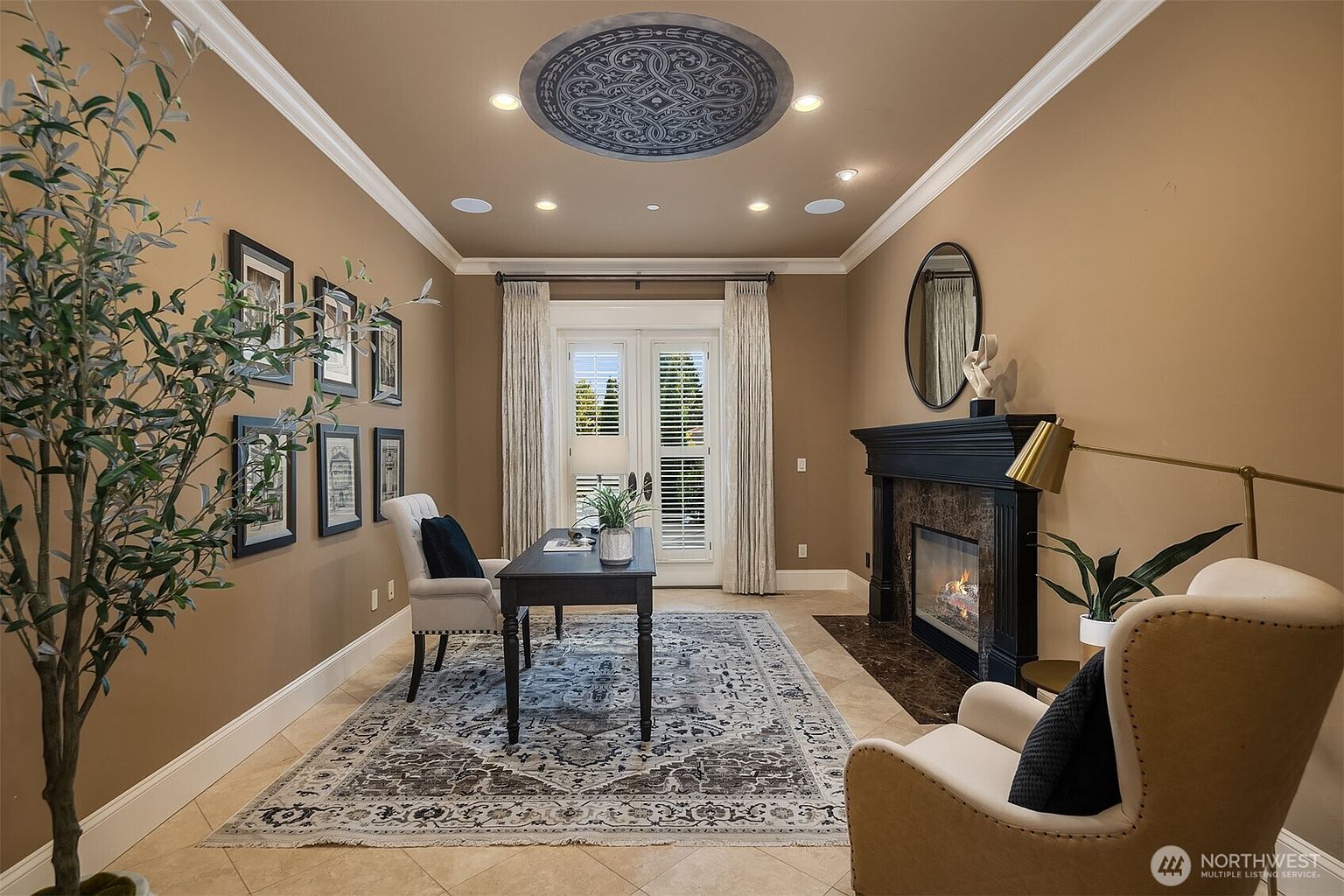 This elegant home office features a sophisticated design with warm tan walls, a decorative ceiling medallion, and a classic fireplace with a dark mantle. A central desk is positioned on a patterned area rug, flanked by a comfortable armchair and a gallery wall of framed art, creating a refined and productive atmosphere. The room is bathed in soft light from recessed fixtures and French doors, offering a serene and professional workspace.
