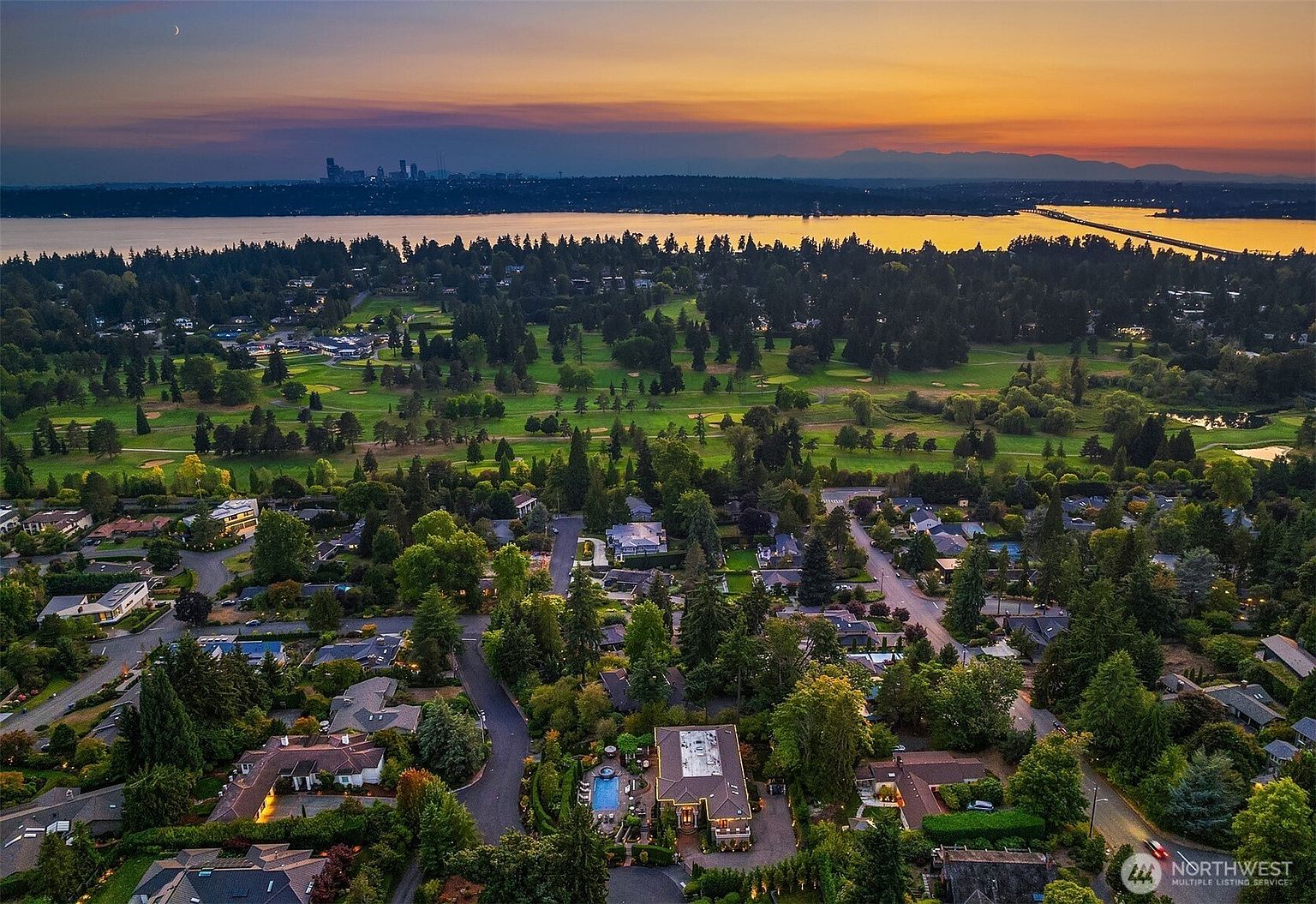 This stunning aerial view captures a luxurious residential neighborhood nestled beside a sprawling golf course, with a breathtaking sunset backdrop over a distant city skyline and water. The perspective highlights the property's prime location, showcasing its private pool, lush landscaping, and proximity to both nature and urban amenities. The golden hour lighting enhances the serene and upscale atmosphere of this expansive suburban setting.