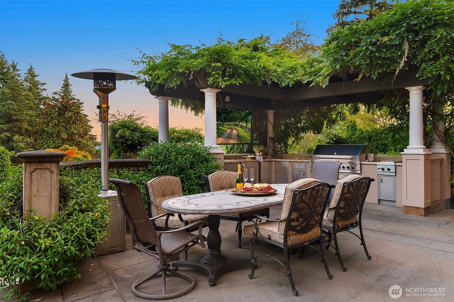 This inviting outdoor patio features a covered dining area complete with a stone-topped table, comfortable cushioned chairs, and a built-in stainless steel grill station. A lush, vine-covered pergola provides shade, while a tall patio heater and a mounted television enhance the space for evening entertainment. The setting offers a serene, elevated view of the surrounding greenery, creating a perfect atmosphere for al fresco dining and relaxation.
