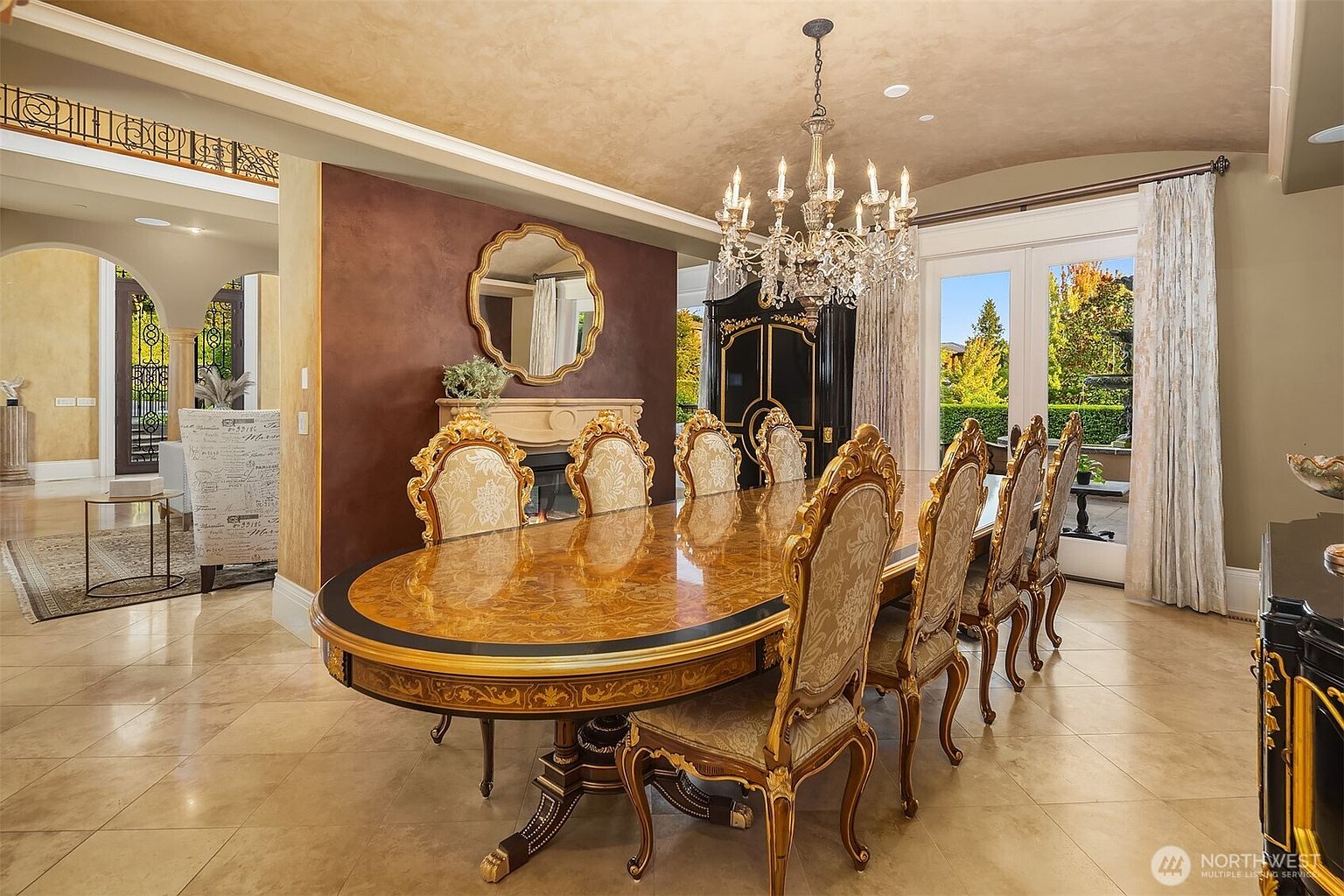 This elegant formal dining room features a grand, ornate wooden dining table surrounded by high-backed, upholstered chairs, set beneath a sparkling crystal chandelier. The room is anchored by a rich, dark-toned accent wall with a decorative mirror above a fireplace, while light-colored tile flooring and large glass doors leading to the outdoors create a bright, airy atmosphere. The overall aesthetic is one of sophisticated, traditional luxury with intricate architectural details.