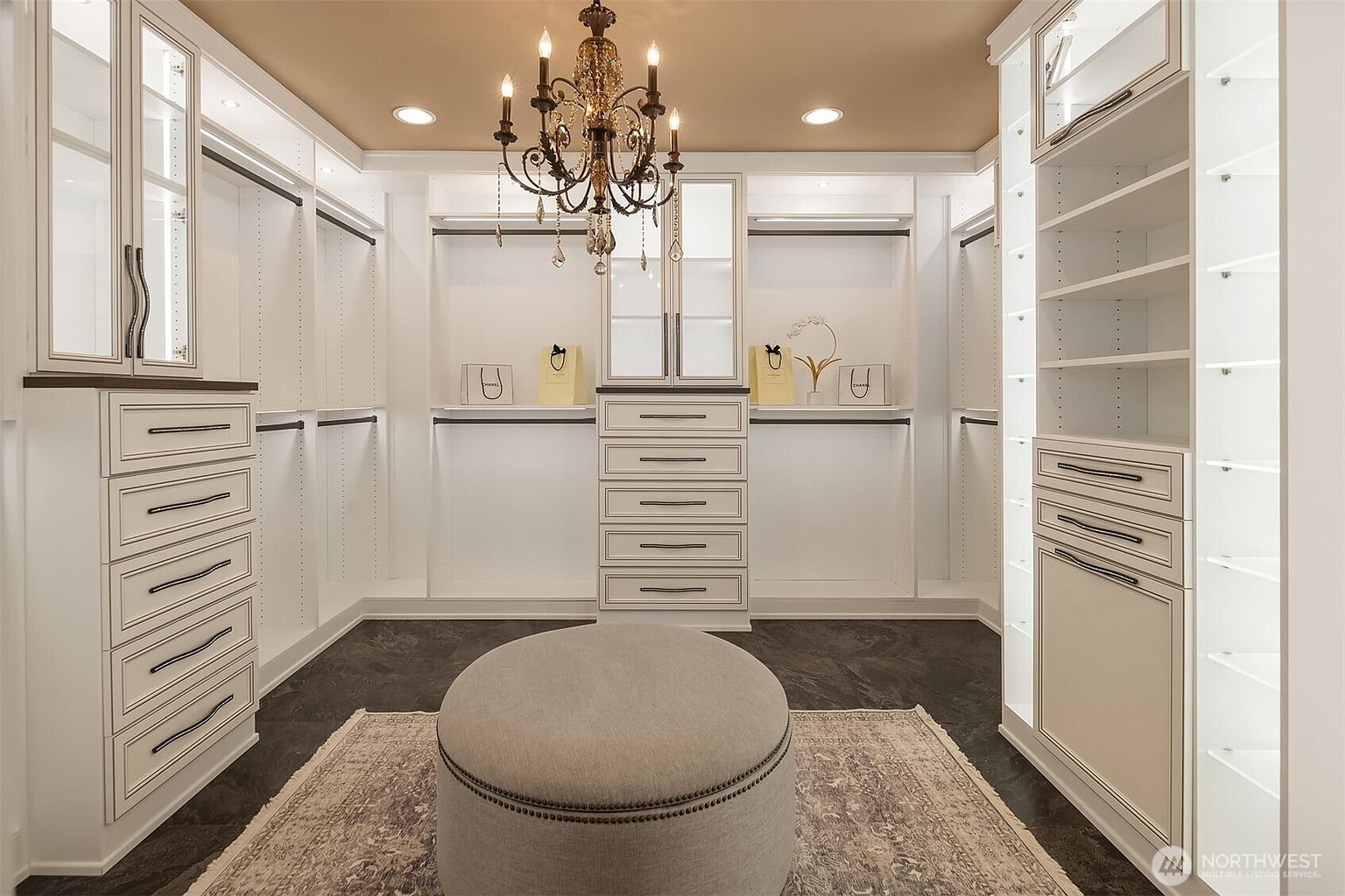 This luxurious walk-in closet features custom white cabinetry with elegant trim, ample hanging space, and integrated shelving for organized storage. A central circular ottoman sits atop a textured area rug, while a sophisticated crystal chandelier hangs from the ceiling, adding a touch of glamour to the space. The dark flooring provides a striking contrast to the bright, clean aesthetic of the storage units.