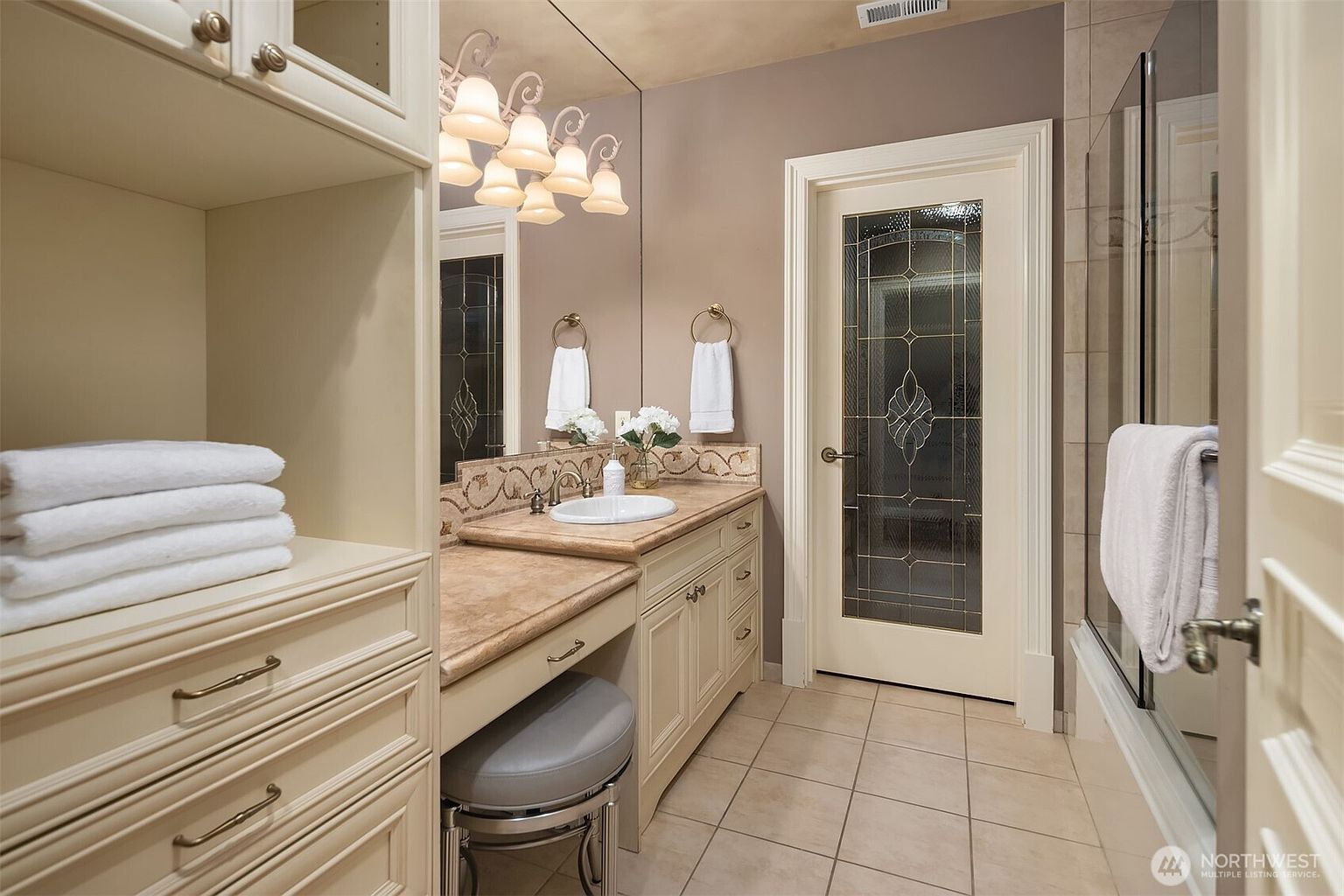 This elegant primary bathroom features a light-toned vanity with a stone countertop, an integrated sink, and a built-in makeup vanity area with a stool. A large mirror reflects the space, which includes a glass-enclosed shower and a decorative glass-paneled door. The room is finished with neutral wall tones and tiled flooring, creating a sophisticated and well-organized atmosphere.