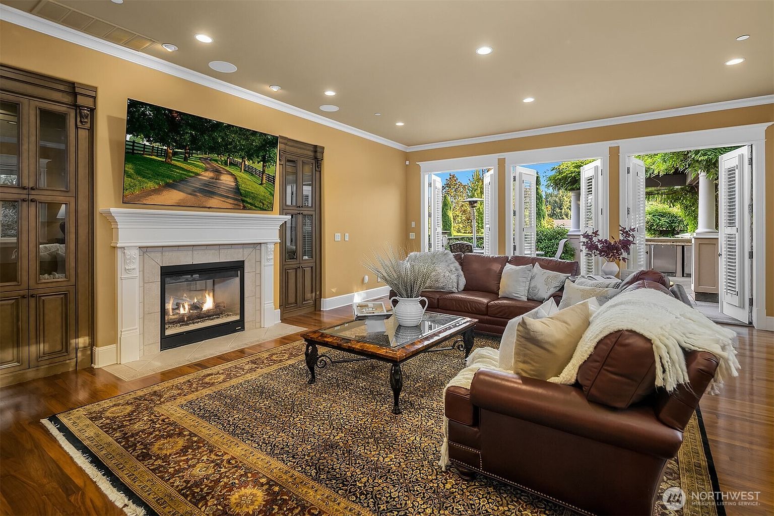 This elegant living room features a warm, inviting atmosphere with rich brown leather sofas and a large, ornate area rug centered on polished hardwood floors. A prominent fireplace with a white mantle serves as the focal point, topped by a large flat-screen television, while built-in wooden cabinetry flanks the fireplace. The room opens up through expansive glass doors to a lush outdoor patio, creating a seamless transition between indoor comfort and outdoor living.