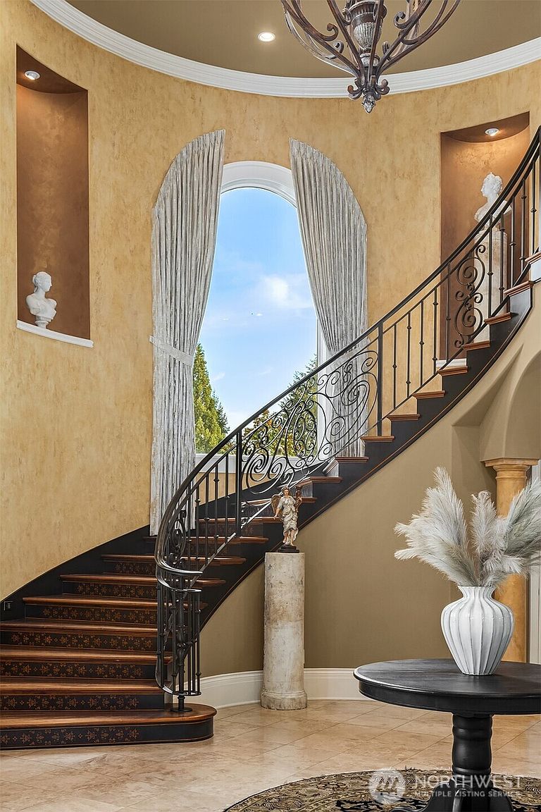 This elegant foyer features a grand, curved staircase with intricate wrought-iron railings and rich wooden treads, serving as the focal point of the space. The walls are finished in a warm, textured faux-gold paint, complemented by arched niches displaying classical busts and a large, light-filled window. A sophisticated chandelier hangs from the ceiling, while a pedestal statue and a circular table with a decorative vase add a touch of refined luxury to the entryway.