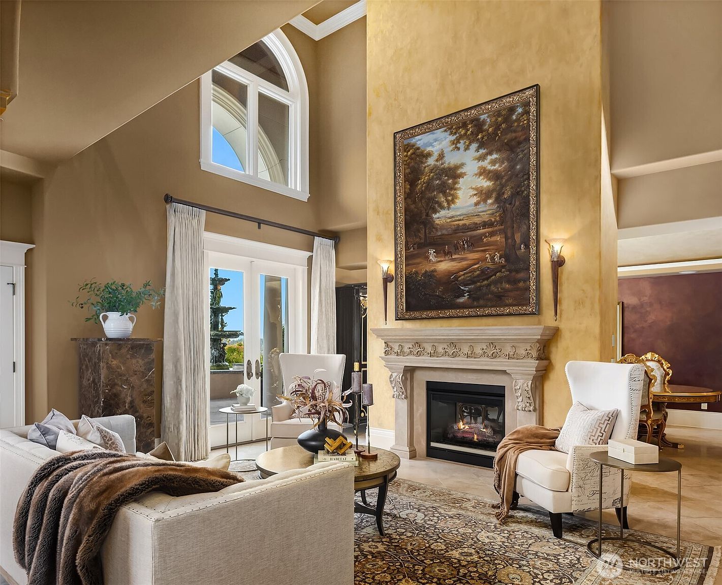 This elegant, high-ceilinged living room features a grand stone fireplace as its focal point, topped with a large, ornate landscape painting. The space is furnished with sophisticated cream-colored wingback chairs and a plush sofa, all arranged on a detailed traditional area rug. Large arched windows and glass doors provide abundant natural light and a view of an outdoor fountain, creating a luxurious and inviting atmosphere.
